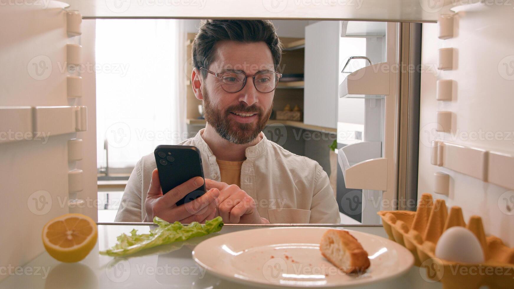 Man hungry guy open empty refrigerator with half lemon, nasty lettuce leaf, one egg, slice of bread order food delivery buying products with mobile phone deliver app point of view from inside fridge photo