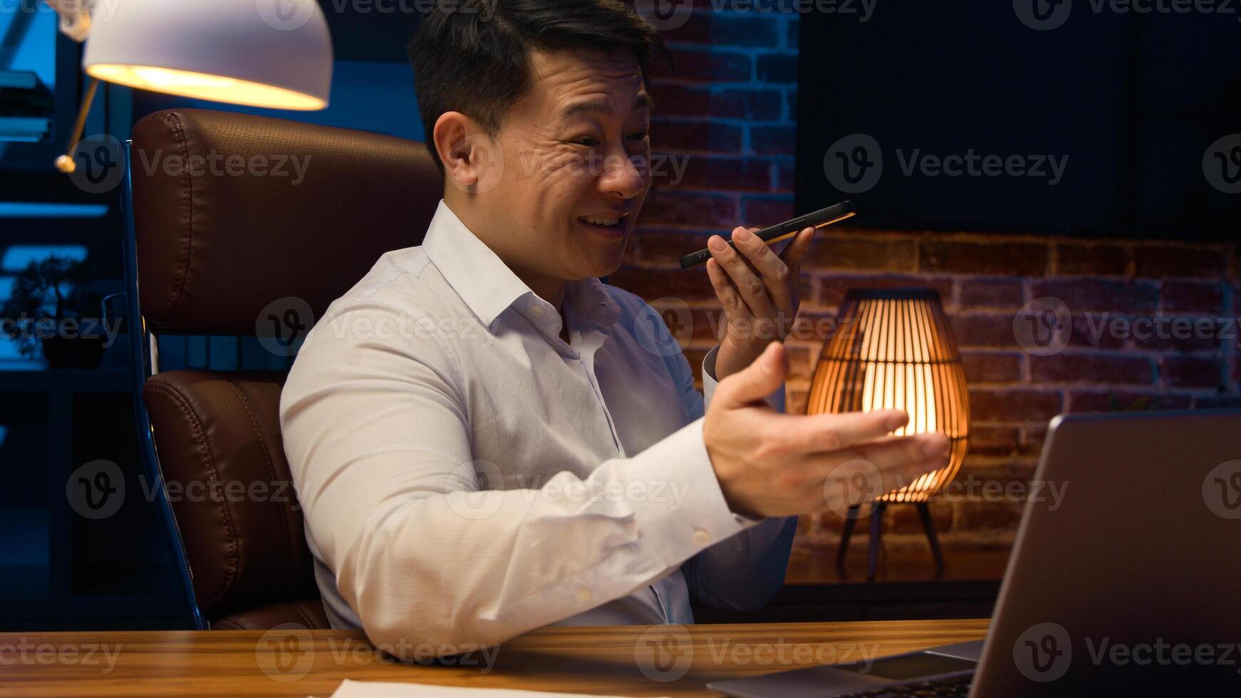 Dissatisfied stressed angry Asian middle-aged office worker at night Korean man businessman laptop error business mistake bad results dictate audio message voicemail mobile phone vocal AI smartphone photo