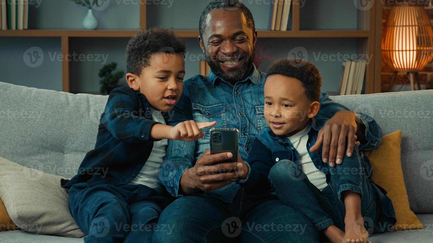 African American happy family father with ethnic sons boys little children siblings kids at home couch sofa using mobile phone together watching funny looking at smartphone app call chat photo