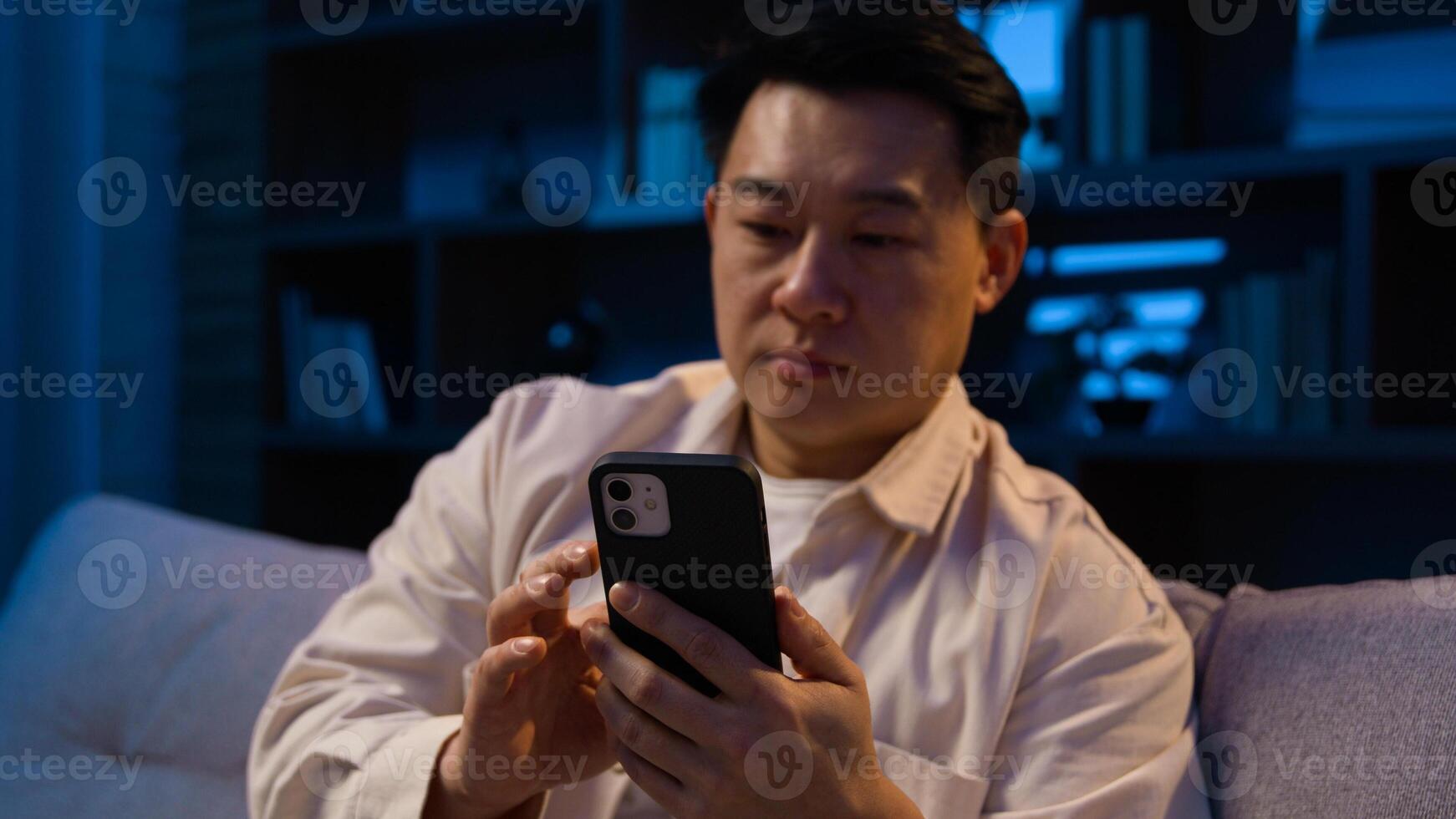 Asian middle-aged man at night evening home at sofa scrolling mobile phone app using gadget addict Korean male with smartphone shopping online social media browsing chatting cellphone at nighttime photo