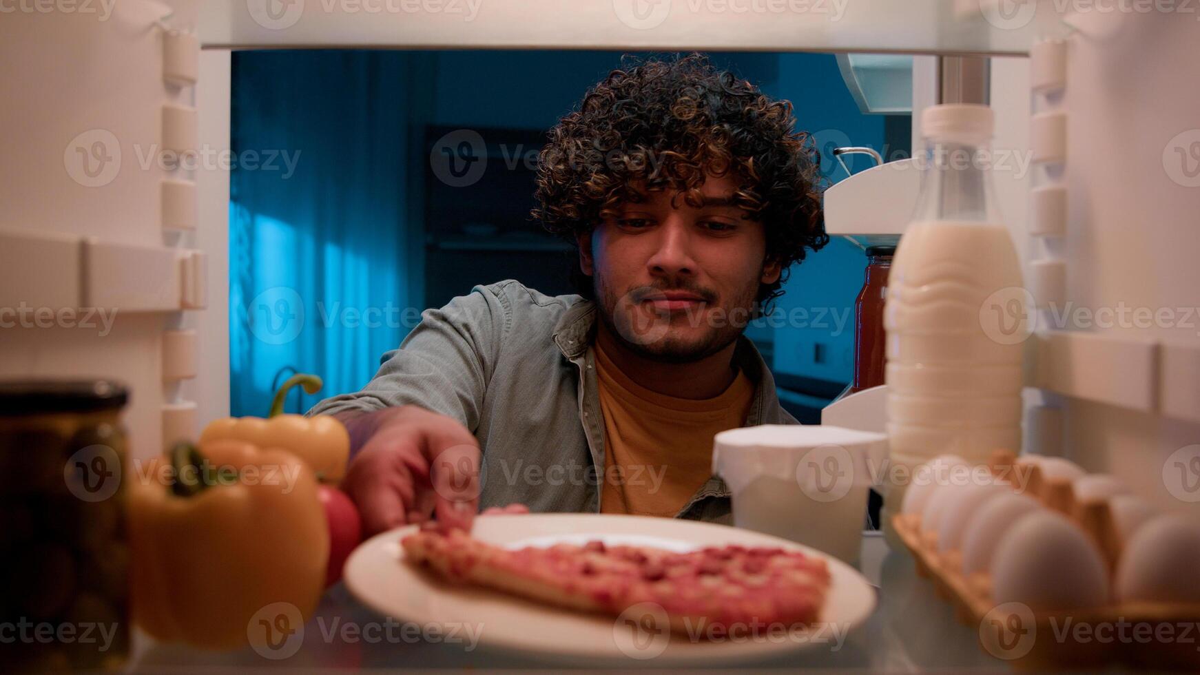 View inside from refrigerator Indian sly man with curly hair open fridge at night evening in kitchen hungry guy male looking around take out slice of pizza overeating at nighttime unhealthy nutrition photo