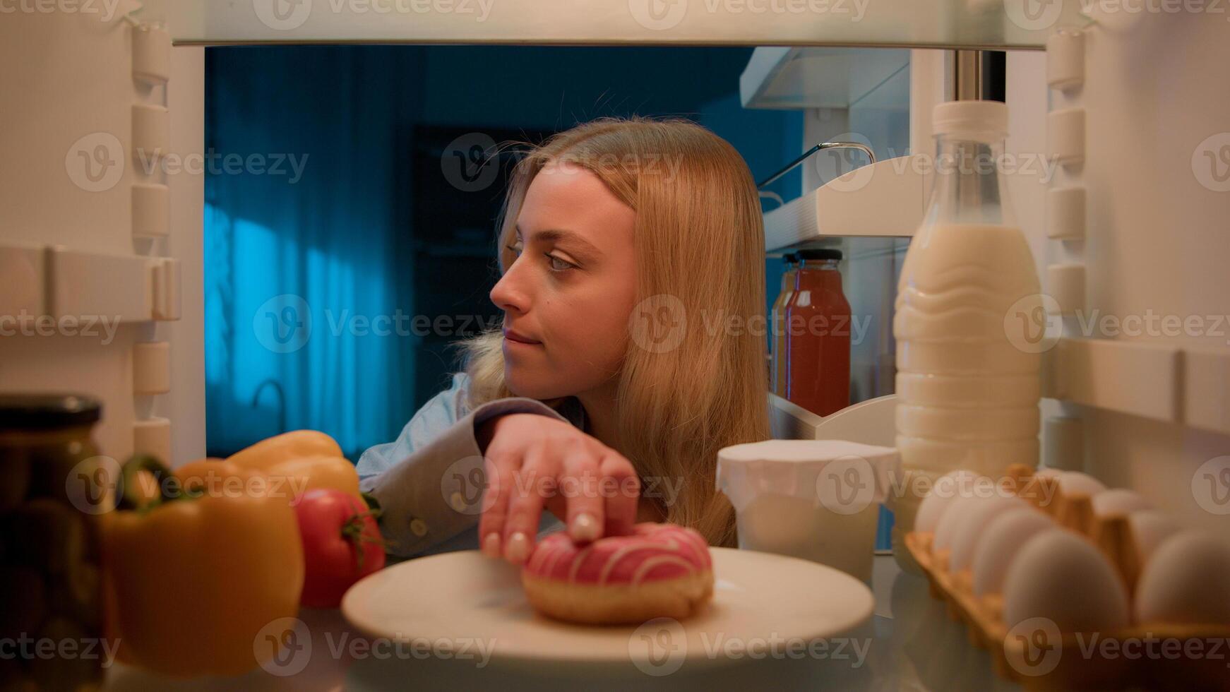 Point of view POV from inside refrigerator hungry Caucasian woman girl at night evening kitchen open fridge take out eating sweet donut unhealthy nutrition obesity diet failure overeating food addict photo