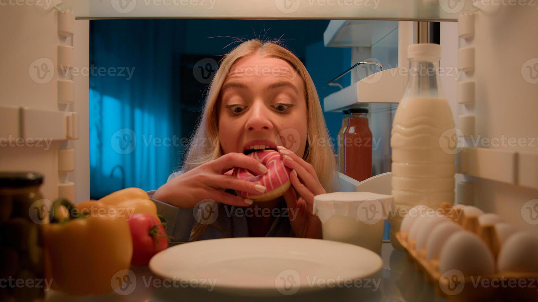 Point of view POV from inside refrigerator hungry Caucasian woman girl at night evening kitchen open fridge take out eating sweet donut unhealthy nutrition obesity diet failure overeating food addict photo