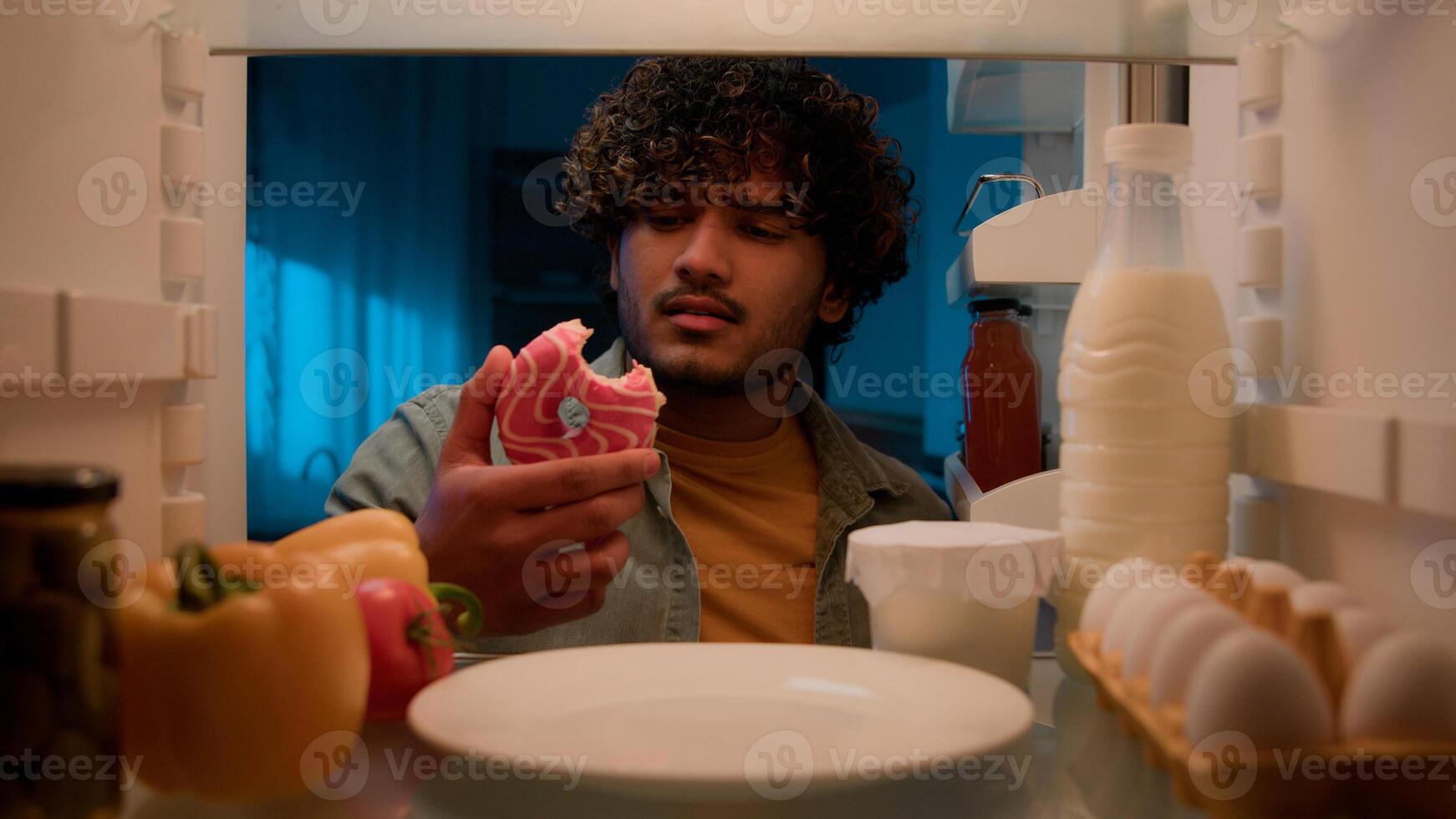 Point of view POV from inside refrigerator hungry Indian man Arabian guy male at night kitchen open fridge want eat take bitten sweet donut smiling condemn bakery unhealthy food nutrition overeating photo