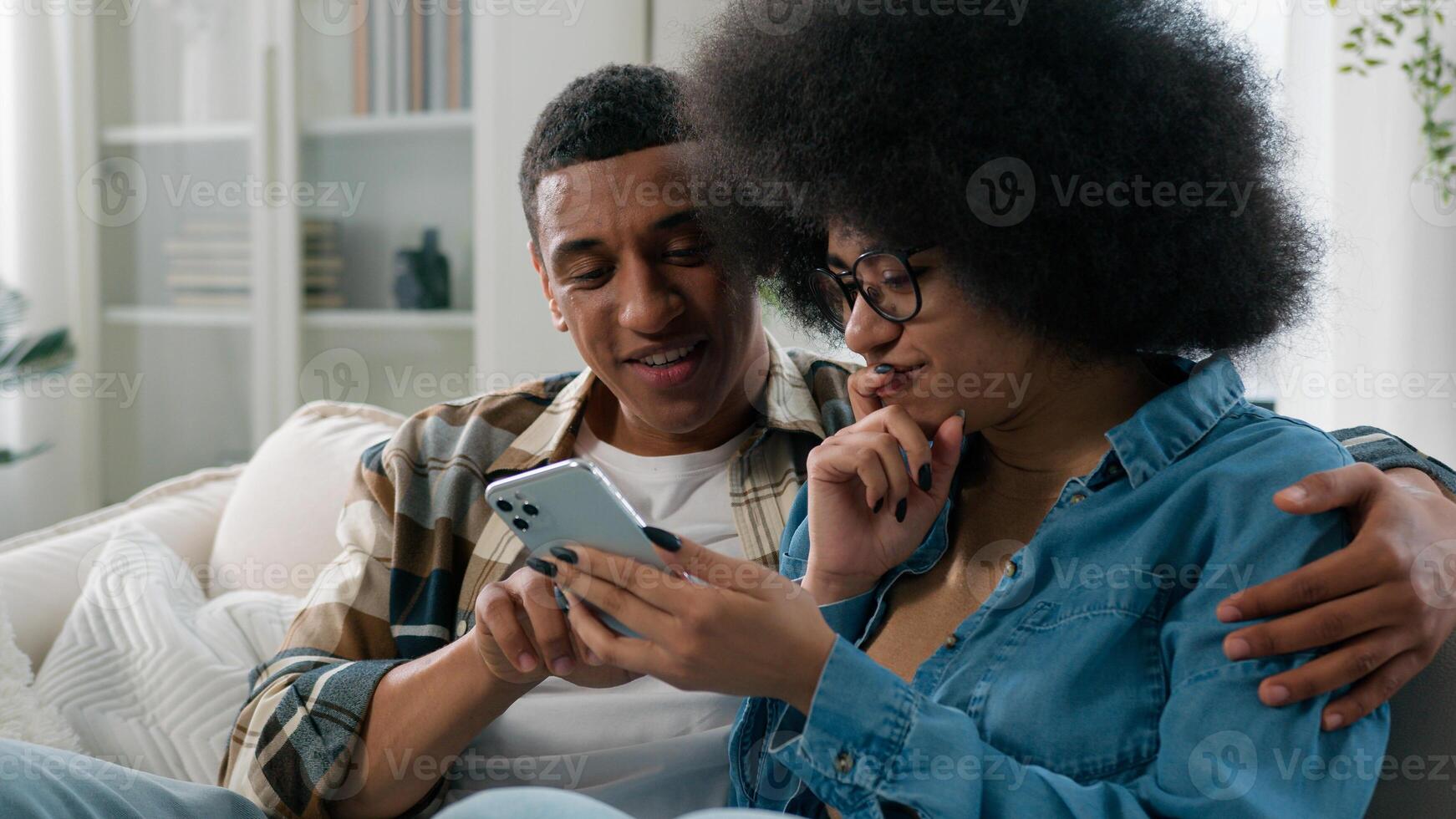 Happy married African American couple man and woman at home couch looking mobile phone shopping website app store internet using smartphone search booking tickets choosing purchase discussing talking photo