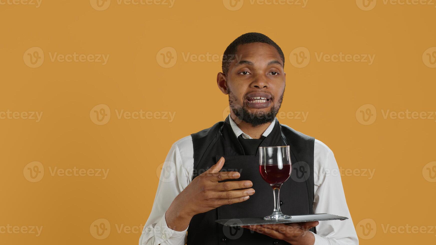 Waiter serving clients with a glass of red wine before dinner, recommending a special drink for customer satisfaction. Employee carrying a tray and serves people with professionalism. Camera A. photo