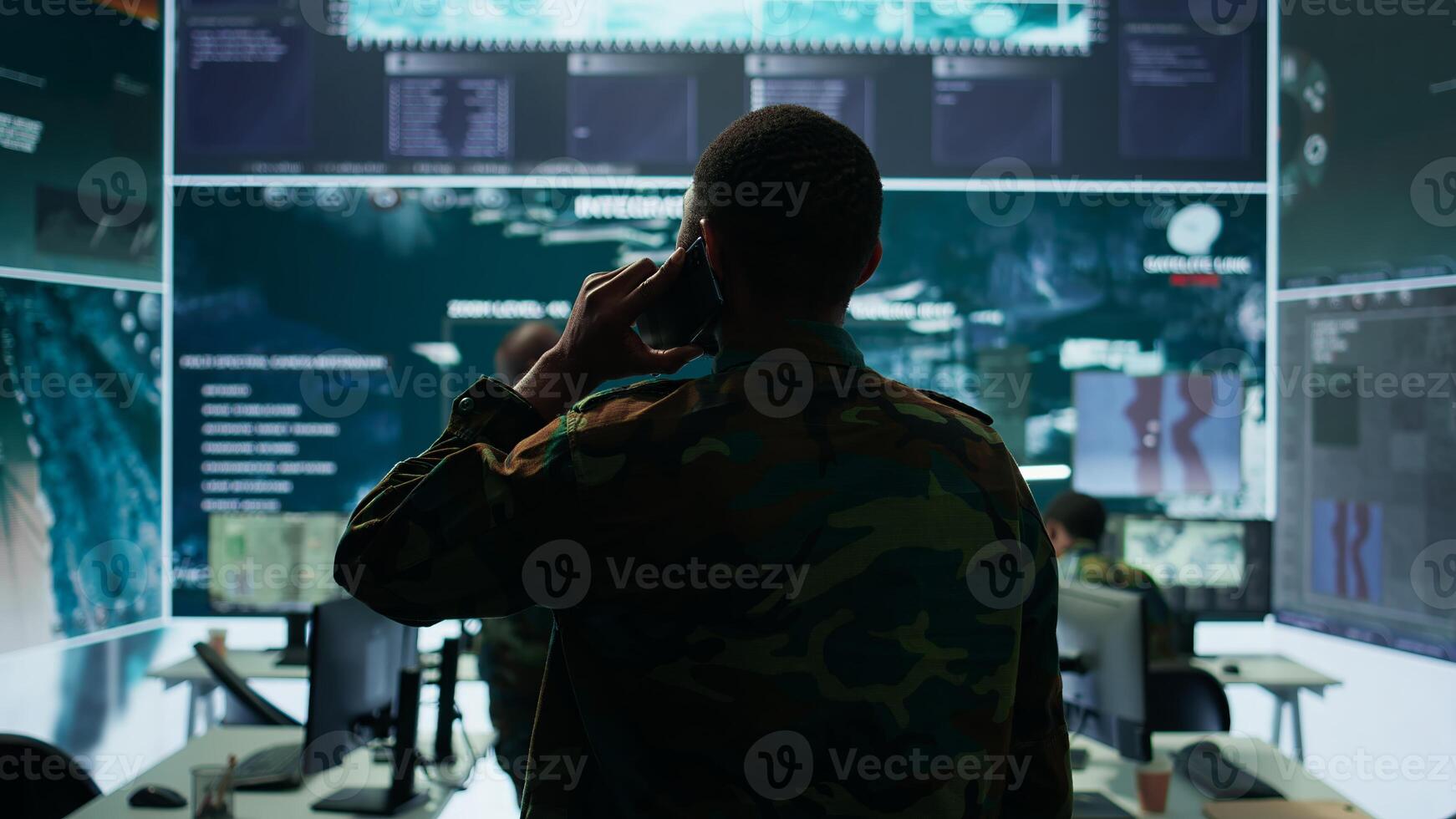 Military man answers phone call in a mission control center, working with advanced technology and satellite imagery to track potential threats and real time data. Intelligence activities. Camera B. photo