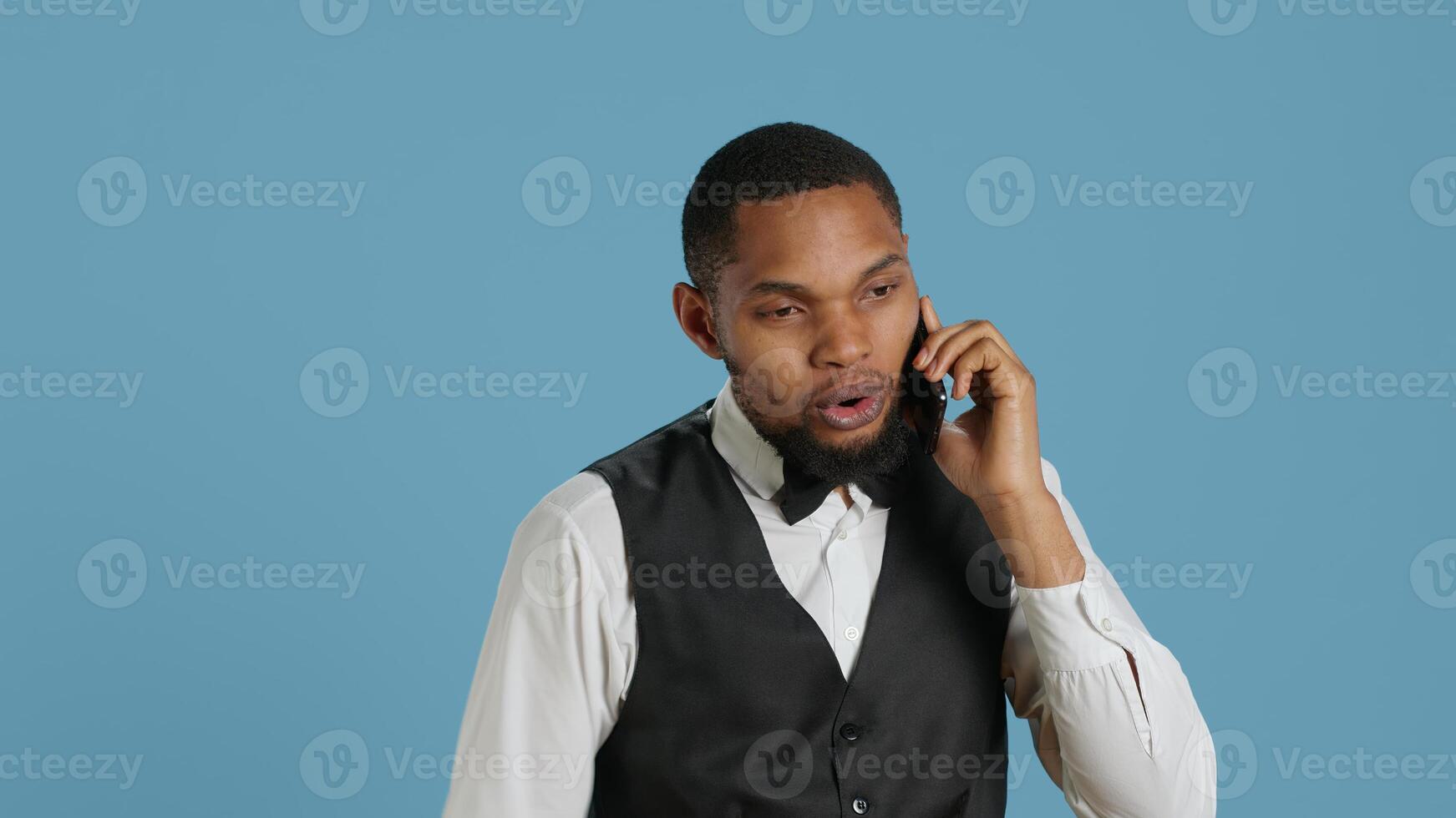 Hotel concierge in uniform makes room reservations on smartphone call, establishing lodging for clients at five star resort. Bellhop explaining amenities to guests in studio. Camera A. photo