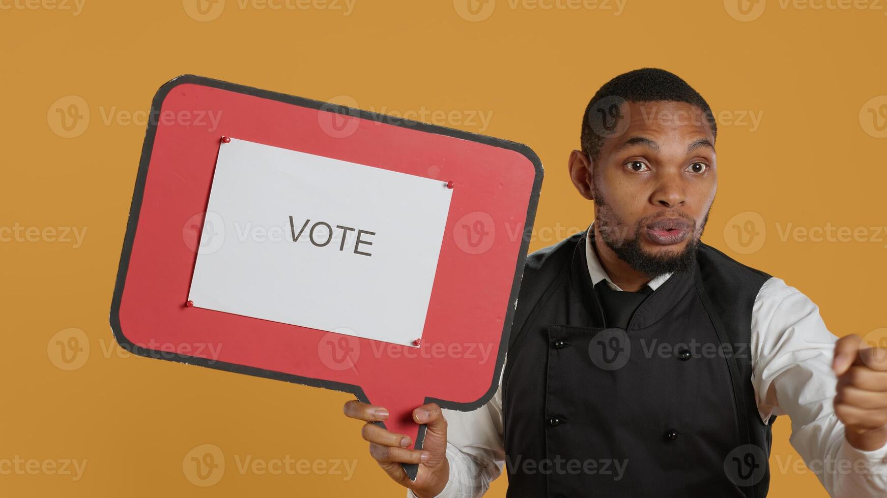 Waiter does an advertisement for politics and showing a speech bubble with the message to vote, standing against yellow background. Restaurant staff holds a cardboard sign. Camera A. photo