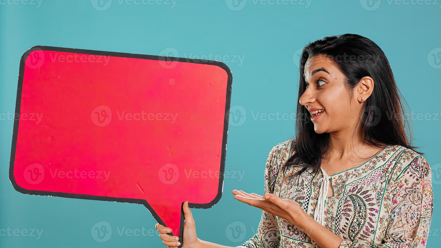 Smiling indian woman holding red speech bubble sign of empty copy space for message. Joyous person presenting thought bubble cardboard used as promotion concept, studio background, camera B photo