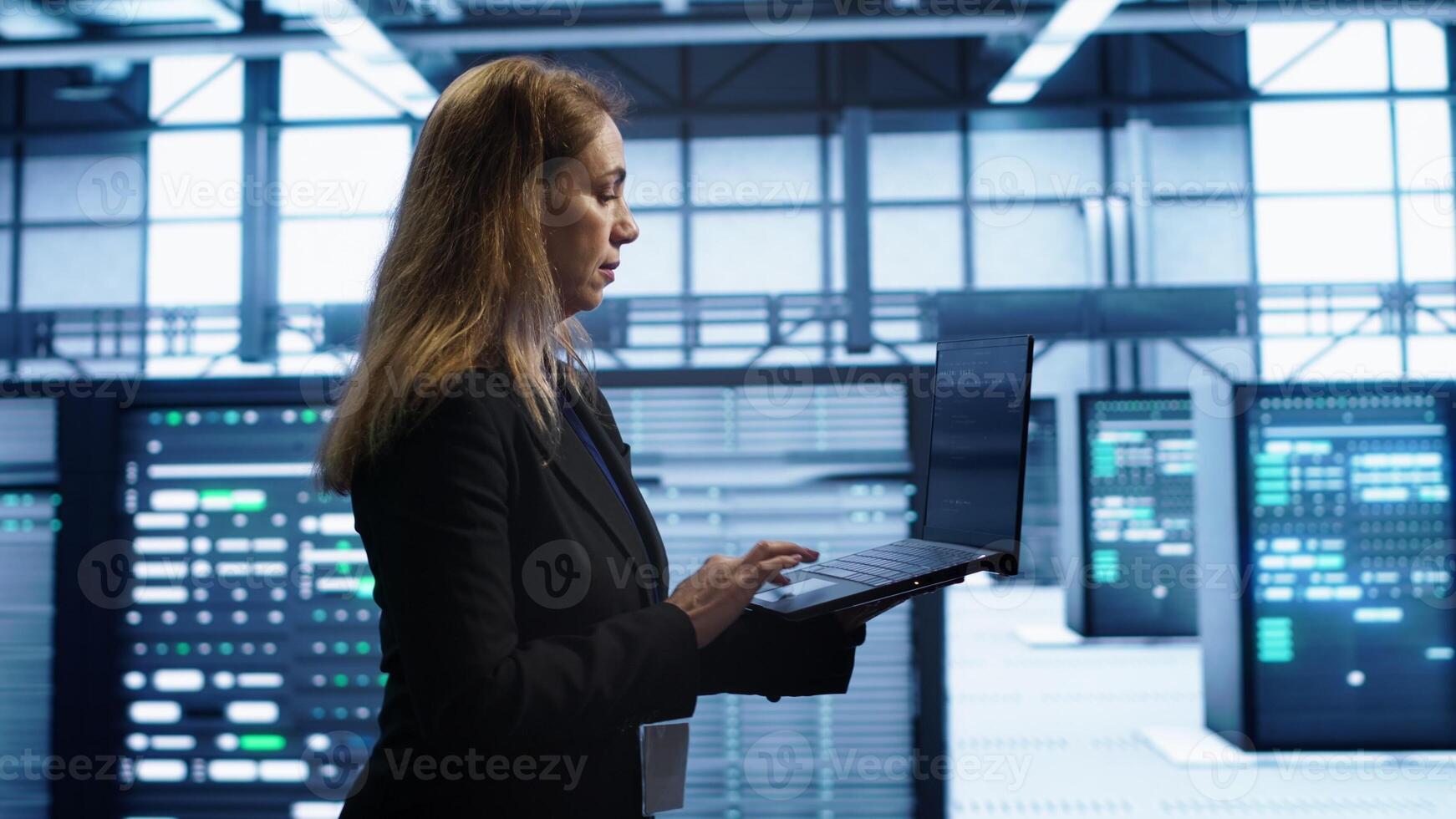 Woman walking in high tech facility, using laptop to patch network issues leading to slow data transfer rates. Employee in data center fixing errors affecting servers performance, camera A photo