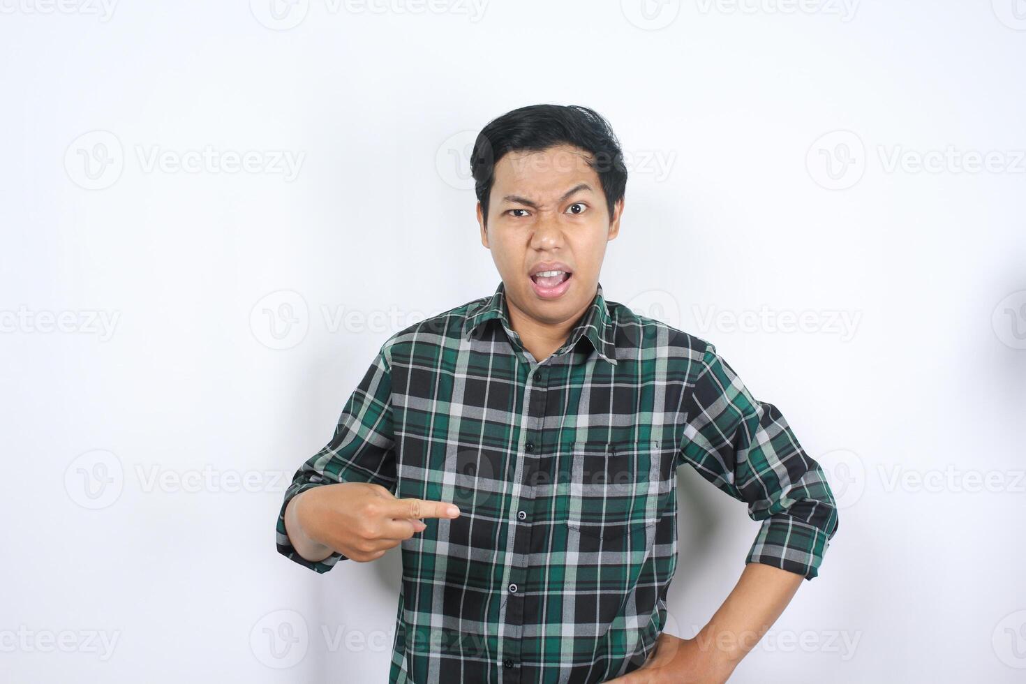 portrait of confused young asian man pointing finger to the middle with shocked and angry face wearing green plaid shirt isolated photo