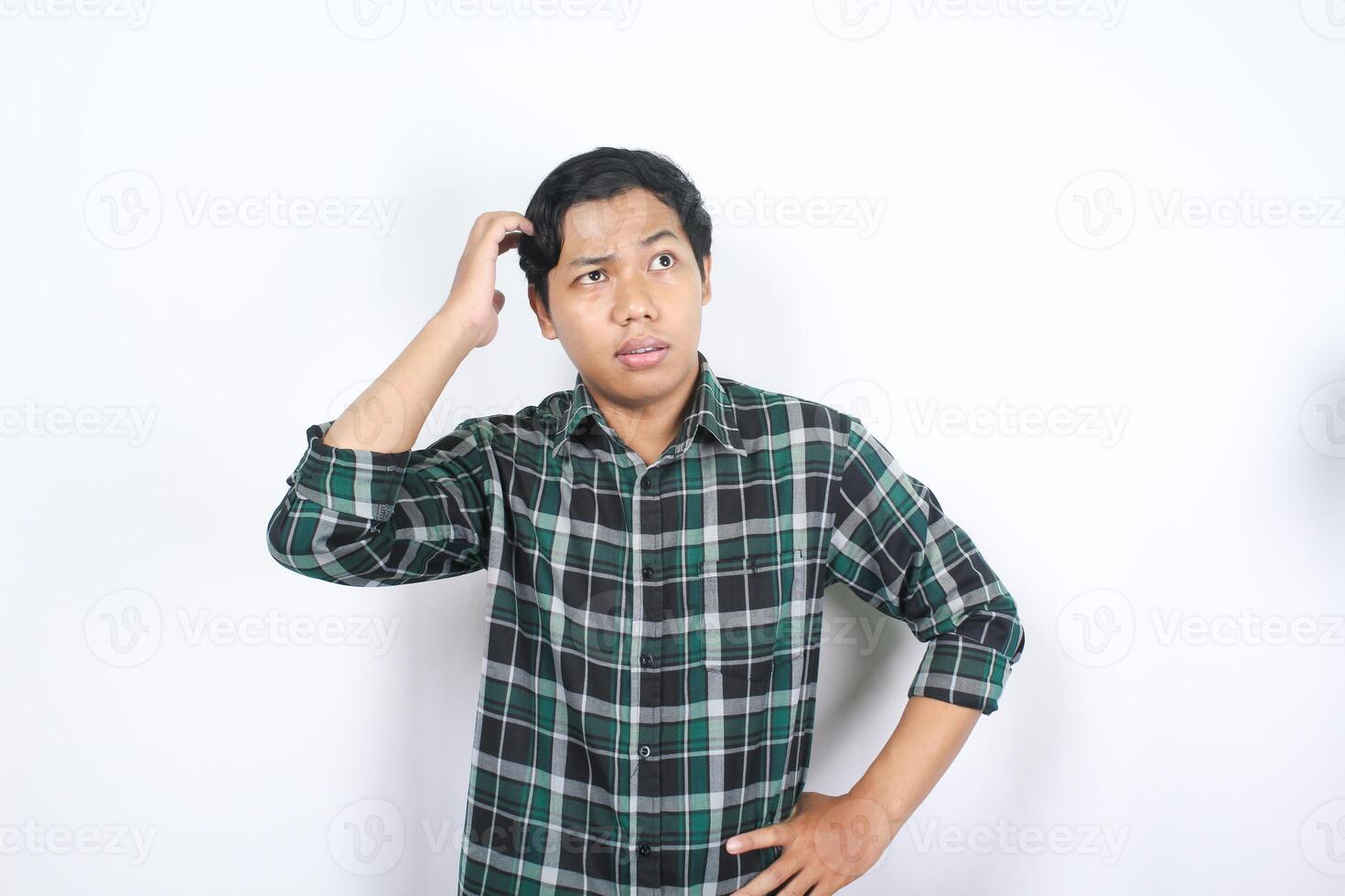 portrait of clueless asian young man wearing casual shirt touching his head and looking corner above with puzzled face isolated on white background photo
