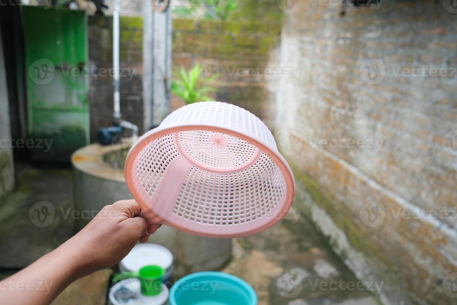 A hand holds a pink plastic strainer, with a blurred background of green plants and stone. photo