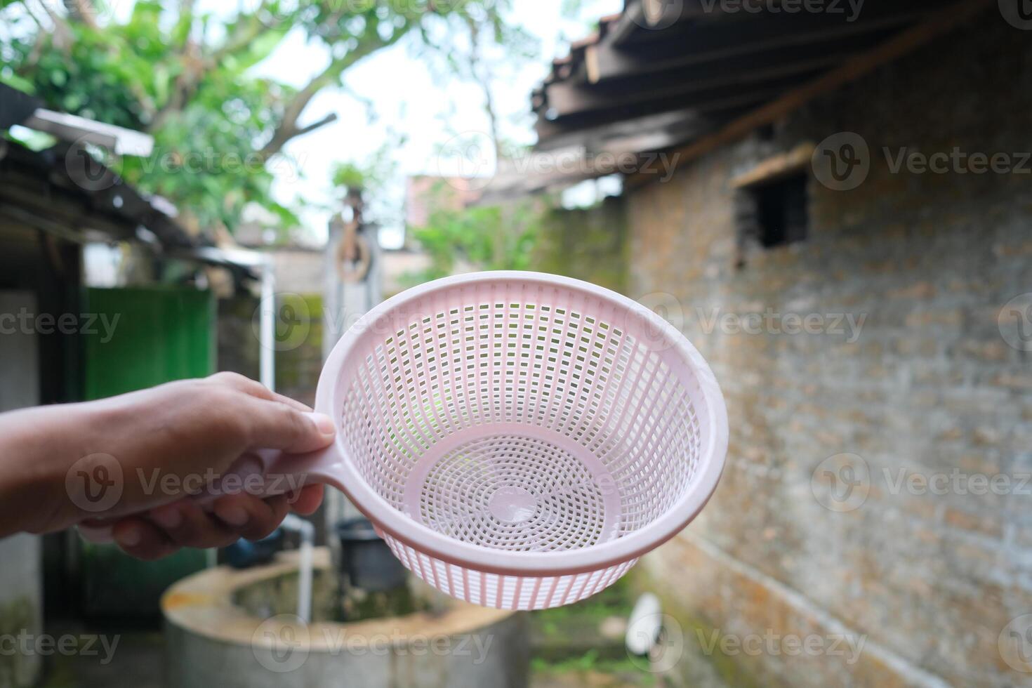 A pink plastic strainer held in a hand, a well in the background. photo