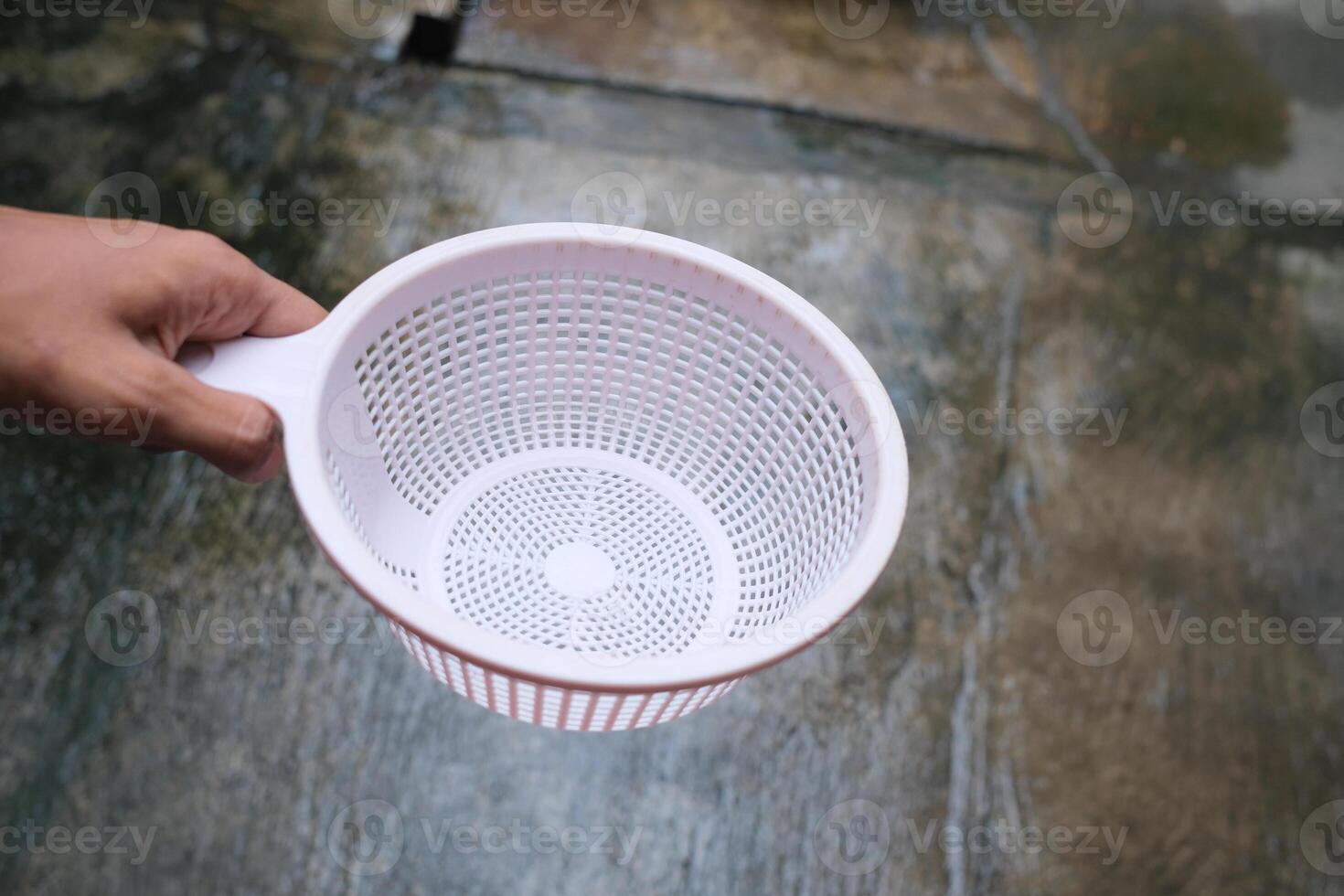 A hand holds a white plastic strainer with a grid pattern against a blurred concrete background. photo