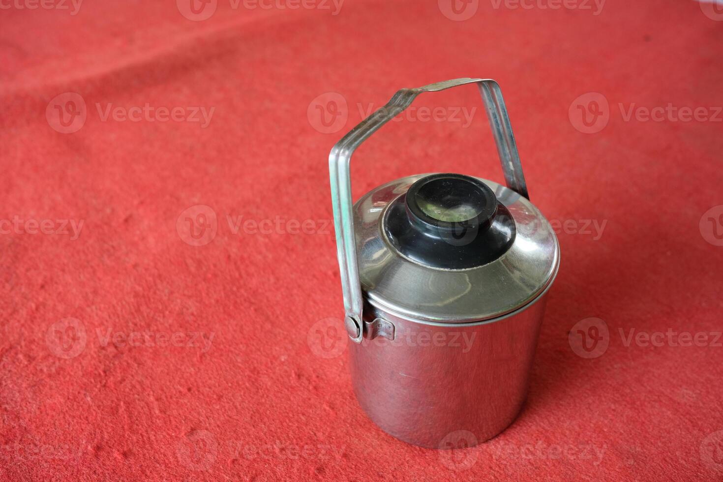 A silver metal container with a black lid and handle sits on a red surface. photo