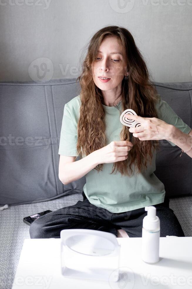 beautiful young woman with long wavy hair doing styling and combing it with a comb, looking in the mirror, sitting on the sofa, at home photo