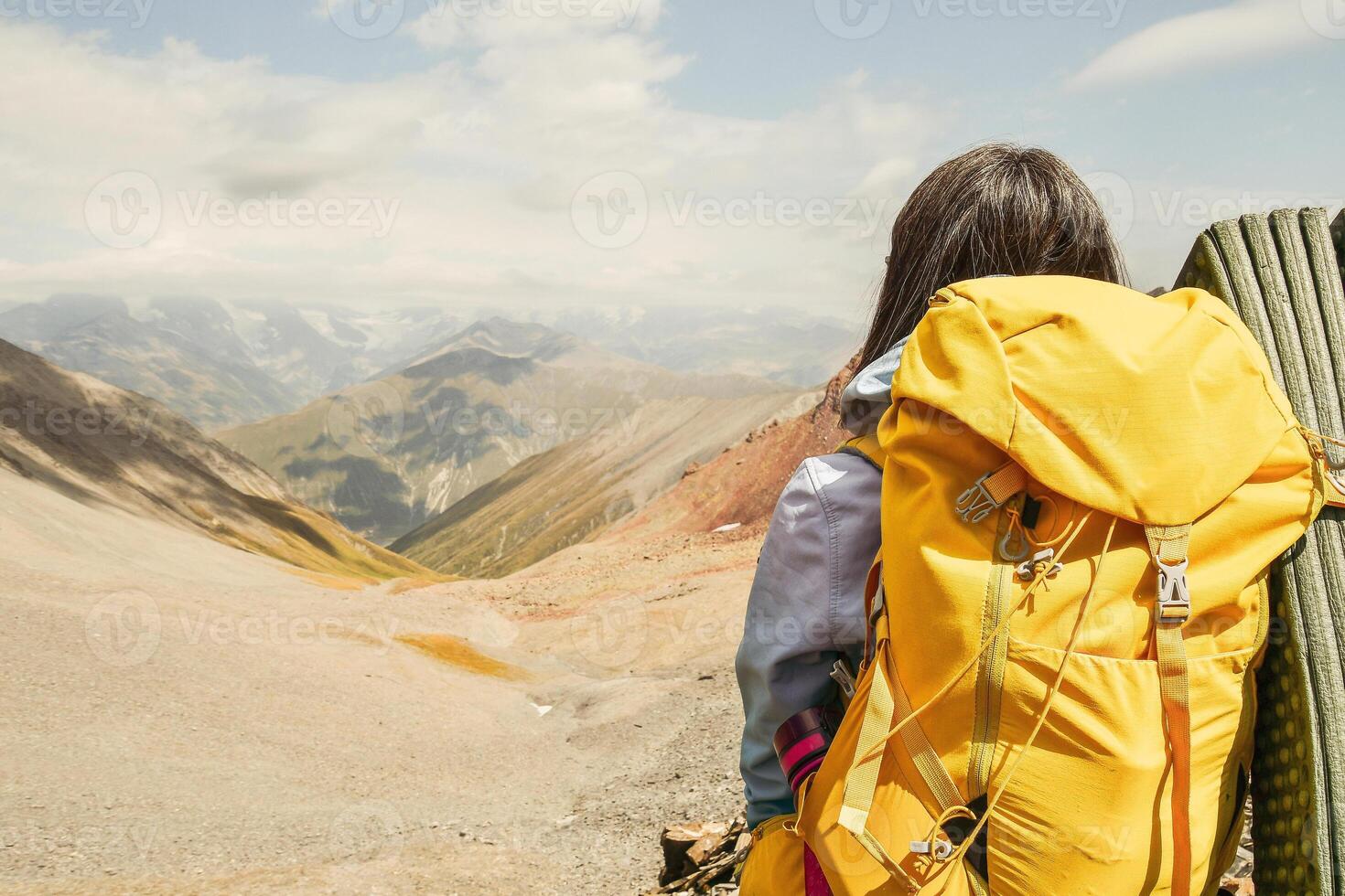 Close up view from the back unrecognizable woman with yellow stylish backpack with flask sticking out of the side pocket. Mock up copy space banner. Woman watch mountains valley in sunny day photo