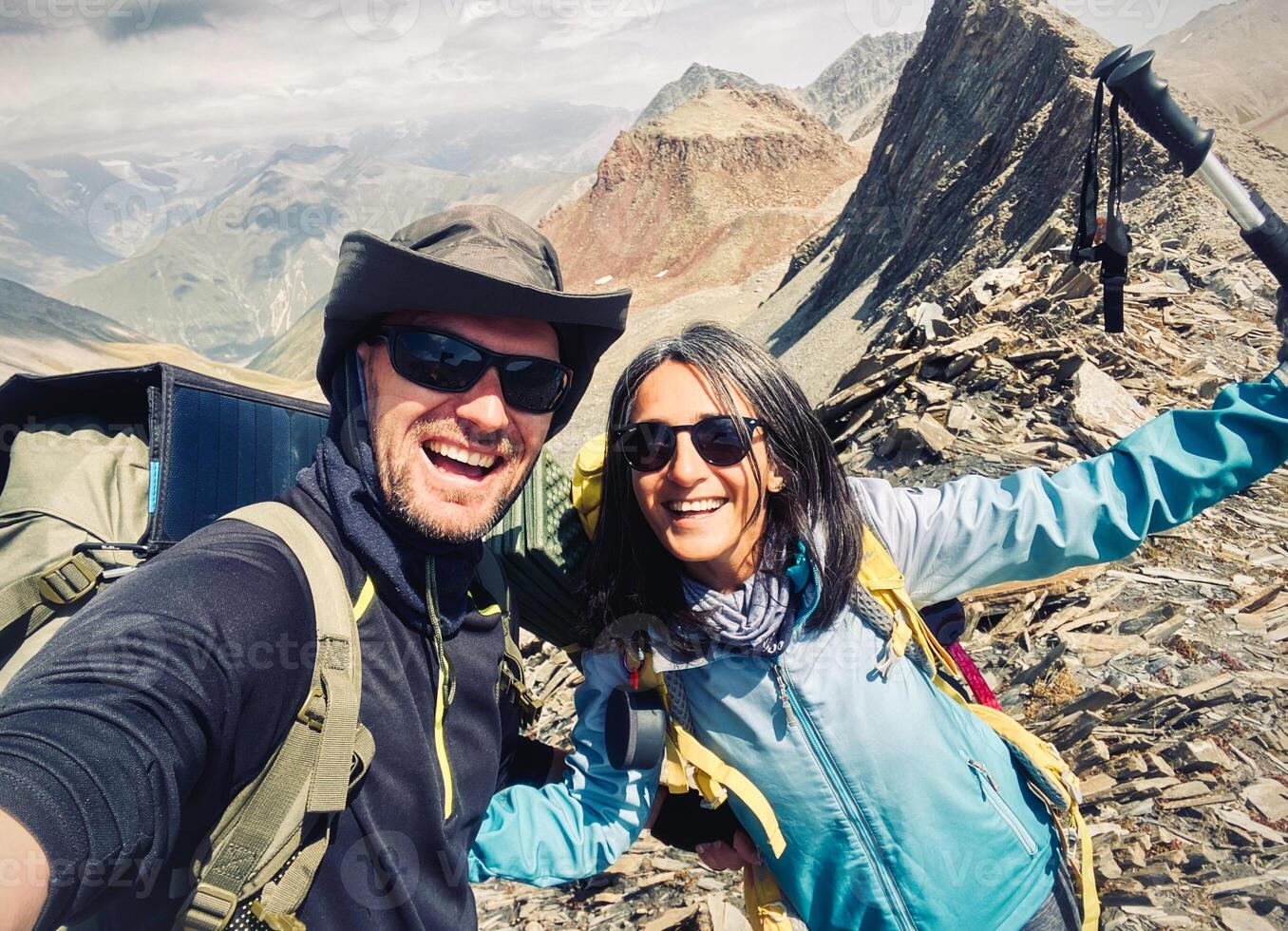 Happy couple of hikers taking selfie picture on top of the mountain - Two travelers with backpack smiling together at camera - Travel blogger influencer photo