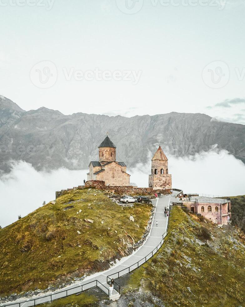 Aerial view Gergeti trinity church after sunset, Georgia. Stepanstminda famous sightseeing landmark on hilltop with misty caucasus mountains background. Tourist visit popular sightseeing attraction photo