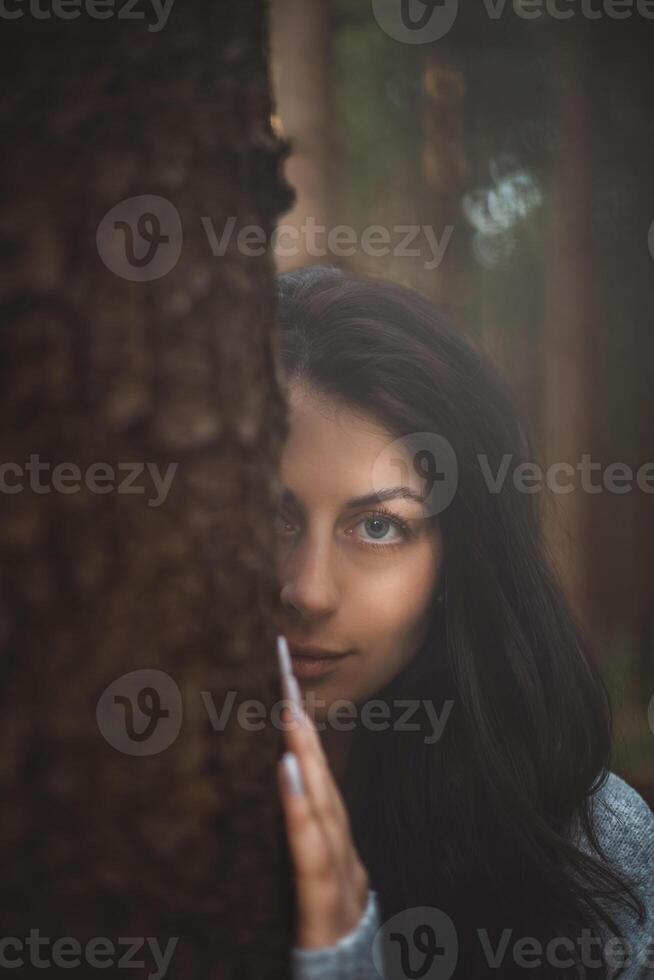 Enigmatic woman with captivating blue eyes peeks from behind a tree in a mystical forest. The image captures the essence of mystery, nature, and serene beauty photo