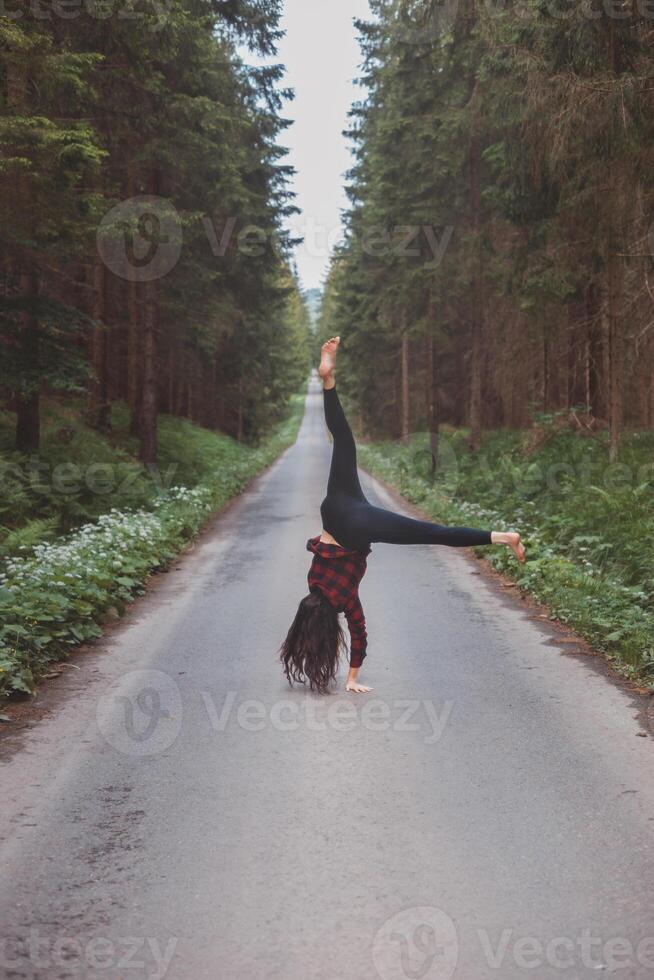 Acrobatic woman in black leggings and plaid shirt performs a cartwheel on a forest path in the middle of a deep forest. Coordination and agility training photo