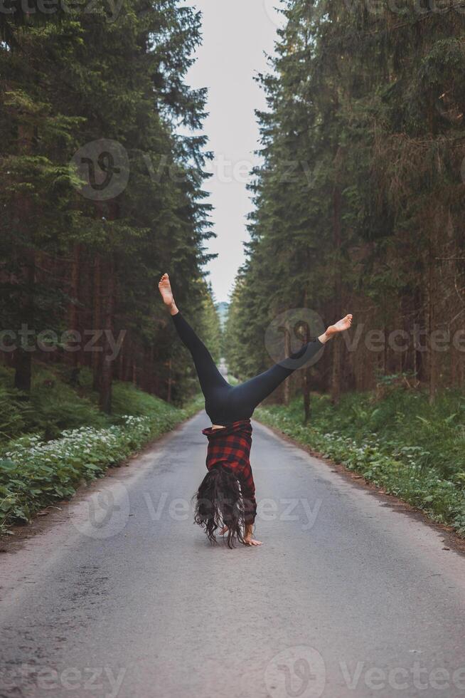 Acrobatic woman in black leggings and plaid shirt performs a cartwheel on a forest path in the middle of a deep forest. Coordination and agility training photo