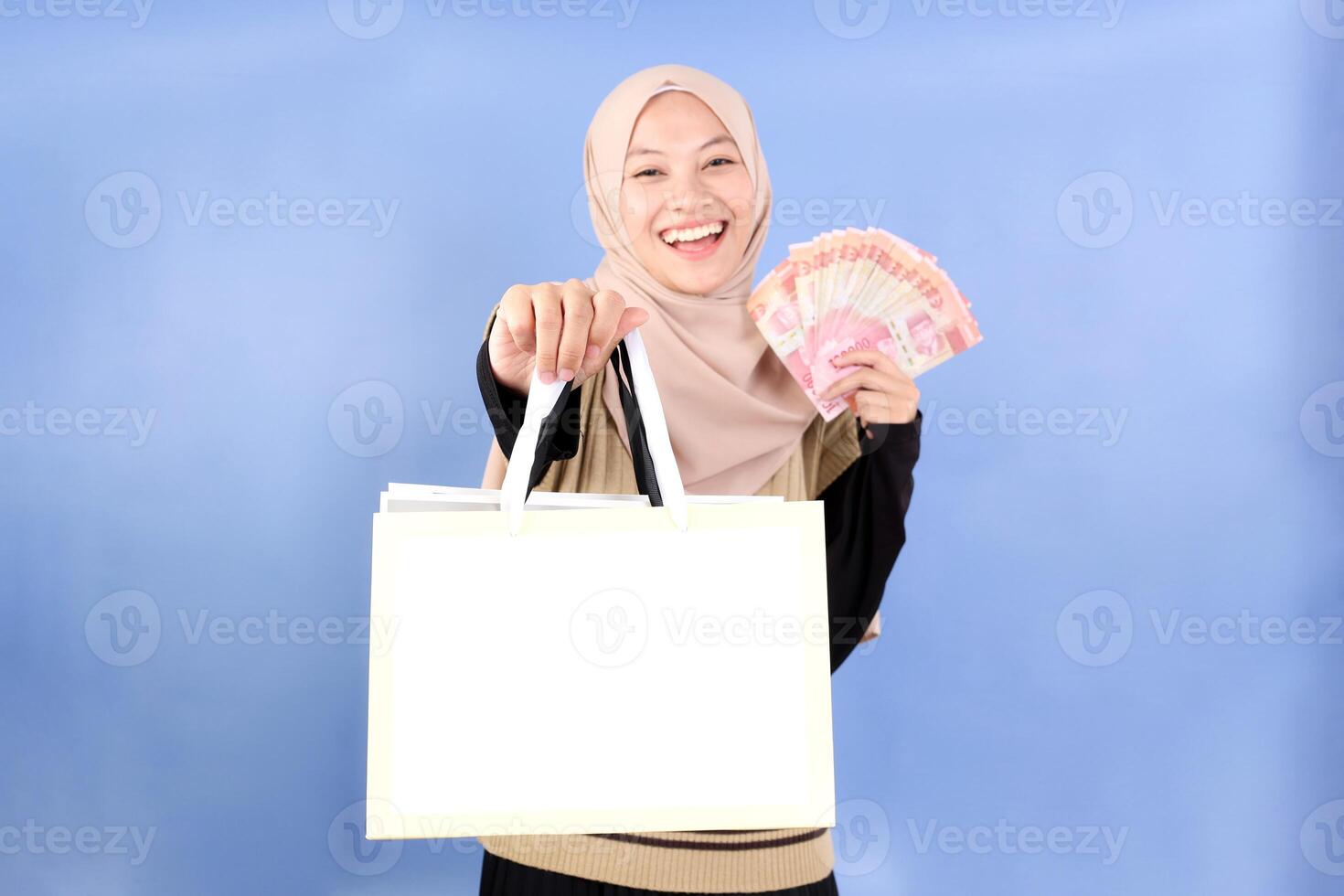 Cheerful Asian Muslim Woman in Hijab Holding Blank Shopping Bag for Mock Up with Rupiah Cash photo