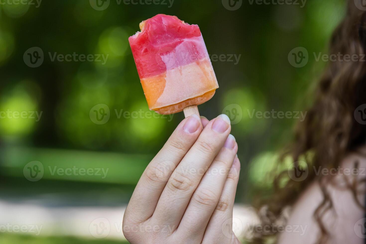 Closeup of the hand holding colorful refreshing partially melted bitten fruit ice cream popsicle on a hot summer day, Selective focus, Burred green outdoor background. Copy space. photo