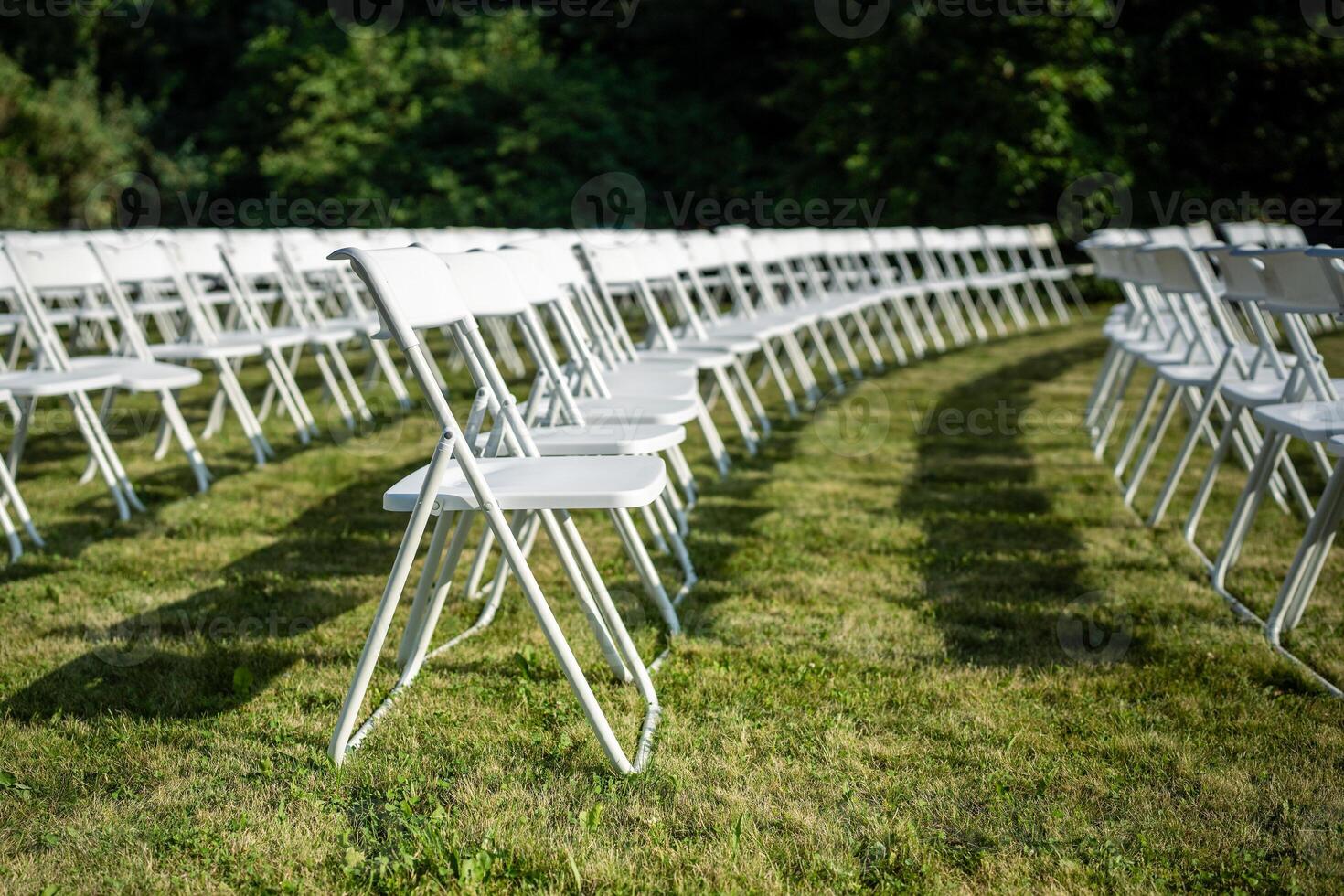 Folding chairs standing in row on green grass, casting shadow on ground. Outdoor party preparation photo