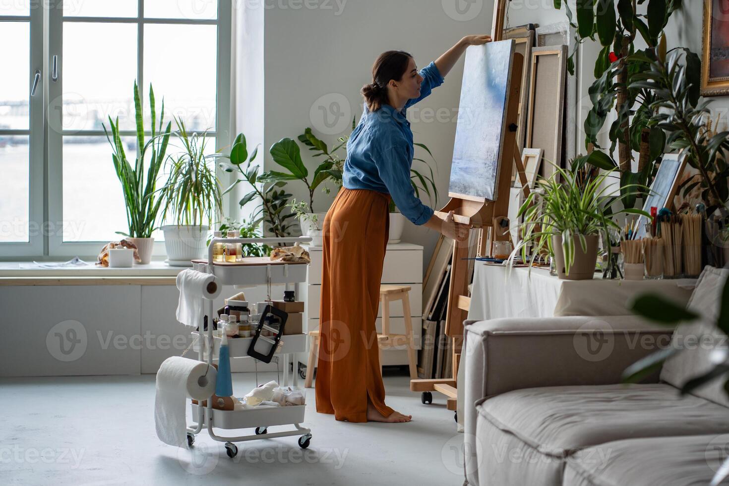 Craftswoman mounting finalized blue painting on easel, taking canvas in hands in radiant art gallery photo