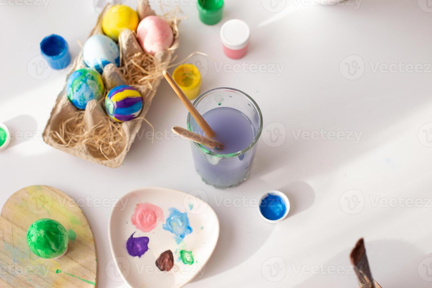 On a white table, in daylight, there are brushes in a glass of water, multi-colored eggs in a tray photo
