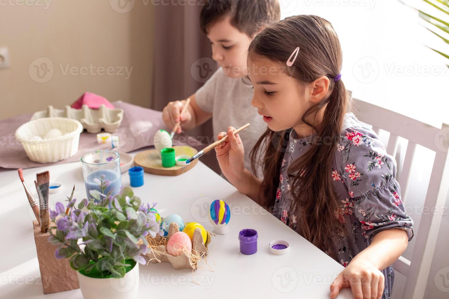 A brunette little girl and boy sits at a white table near the window, paints eggs photo