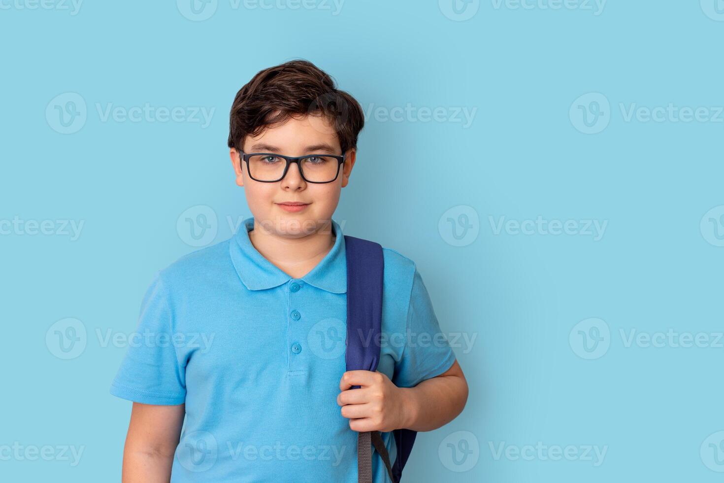 A smiling schoolboy in glasses, with backpack, on blue background, look at camera photo