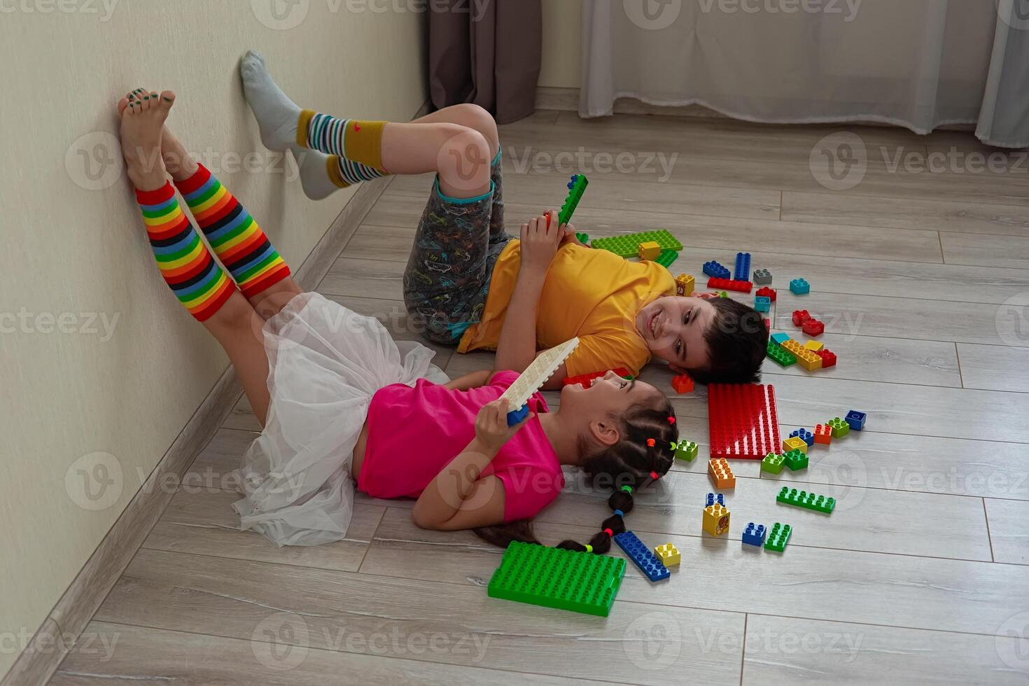 A boy and a little girl, lie on the floor, on their backs, play merrily with a multicolored plastic construction set photo