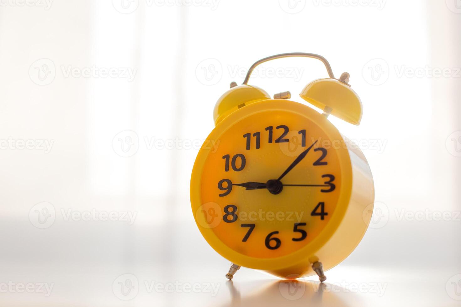 A yellow alarm clock stands on a white table by the window with white curtains photo