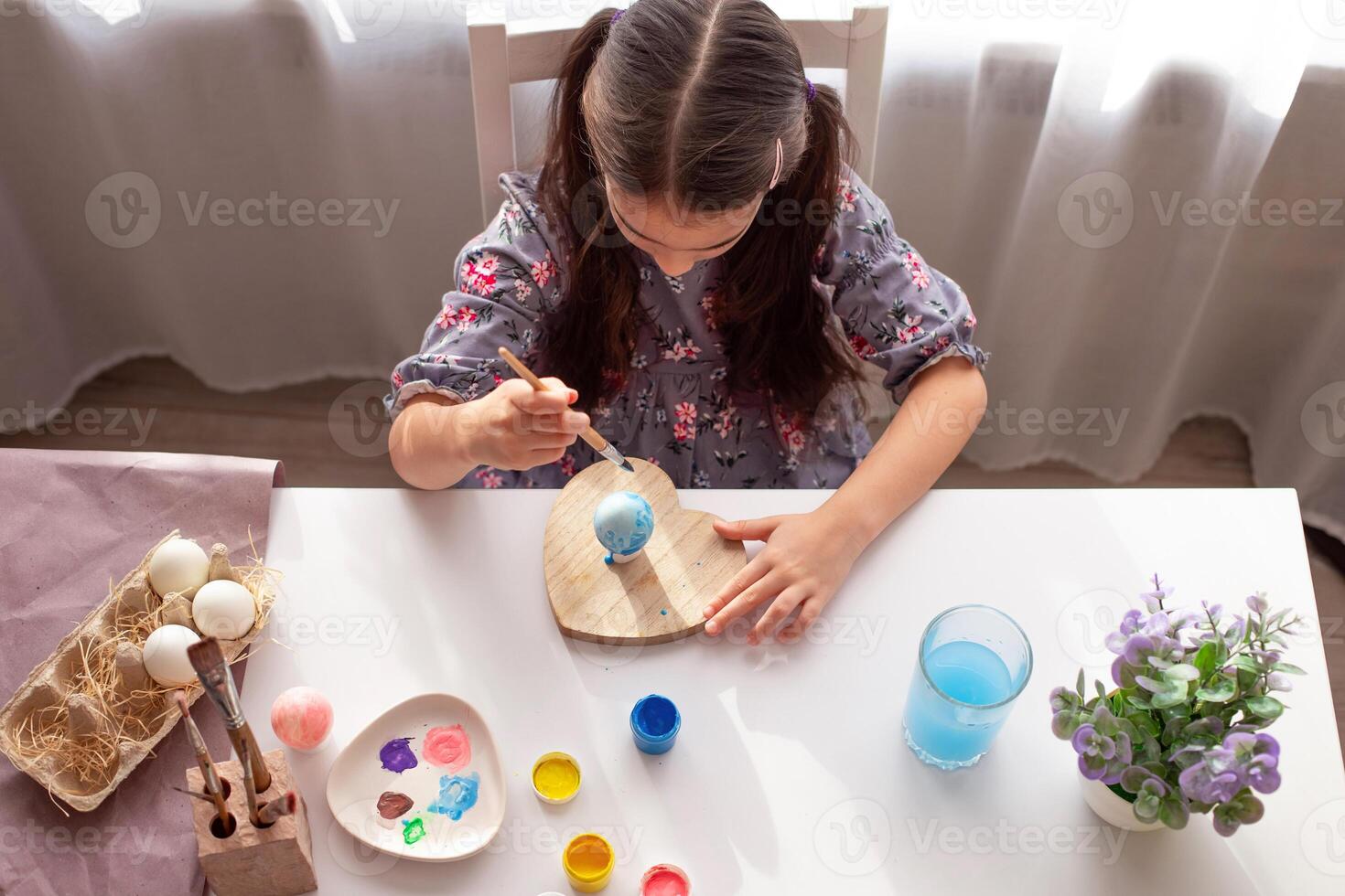 A little girl sits at a white table near the window, paints eggs with a brush. photo