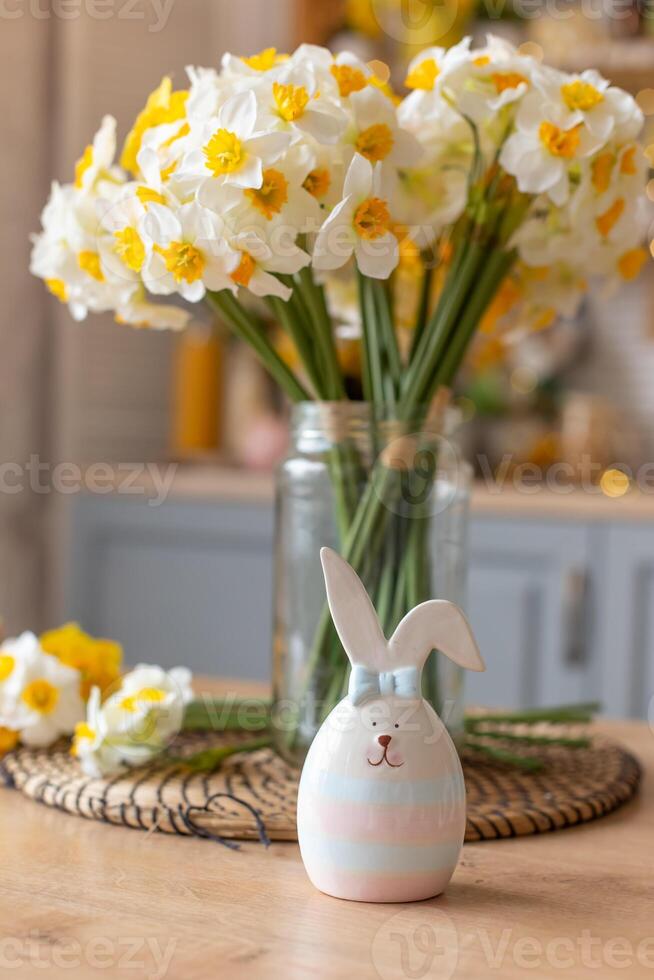 A ceramic figurine of a cute striped hare stands on the kitchen table next to a glass vase with daffodil flowers. photo