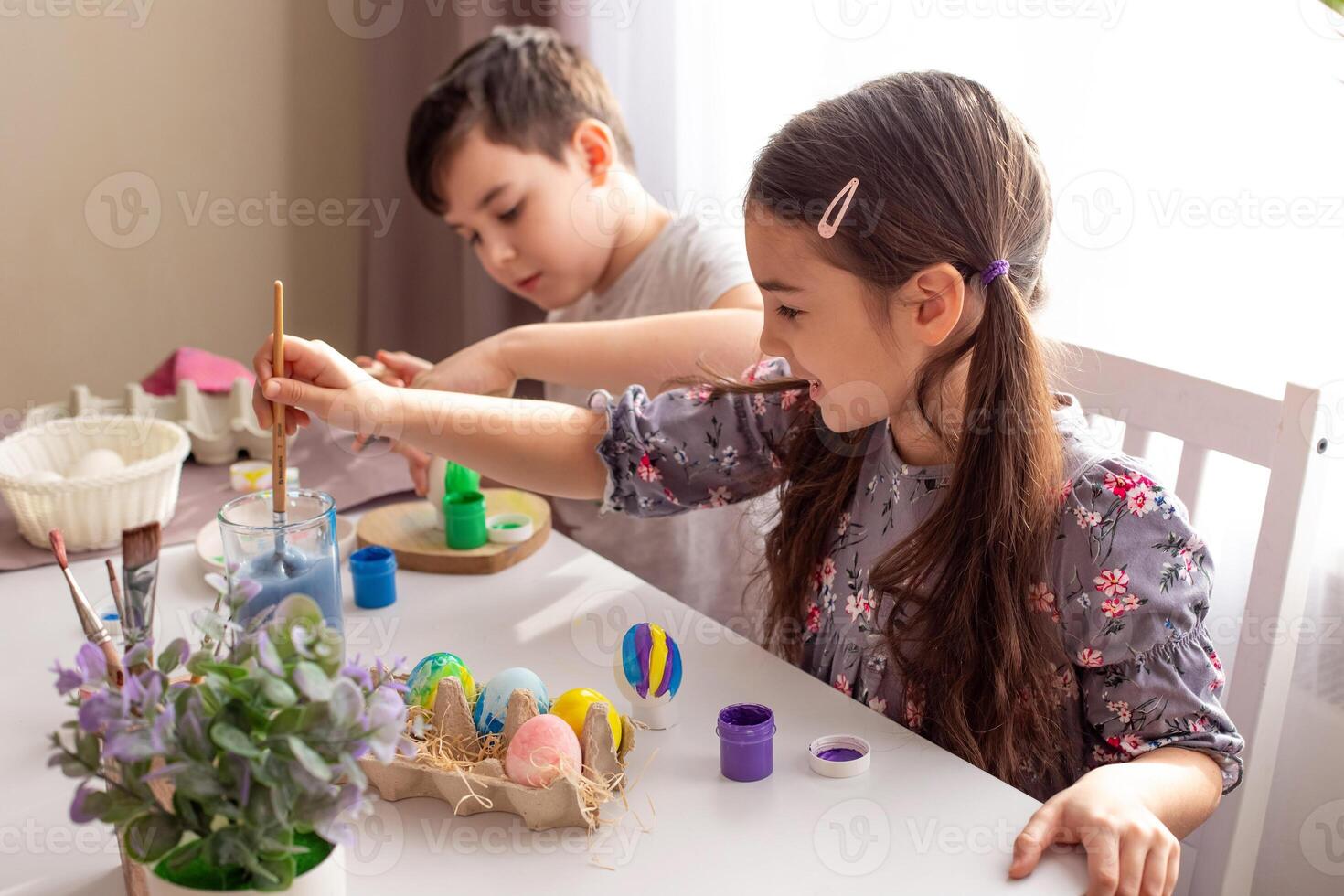 A cute little girl and boy sits at a white table near the window, paints eggs photo