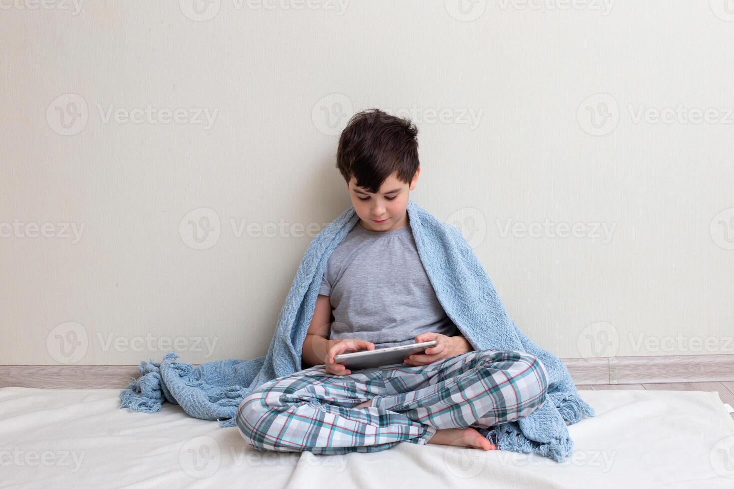 A brunette boy, a teenager in gray pajamas, on the floor, smiles, uses a digital tablet photo