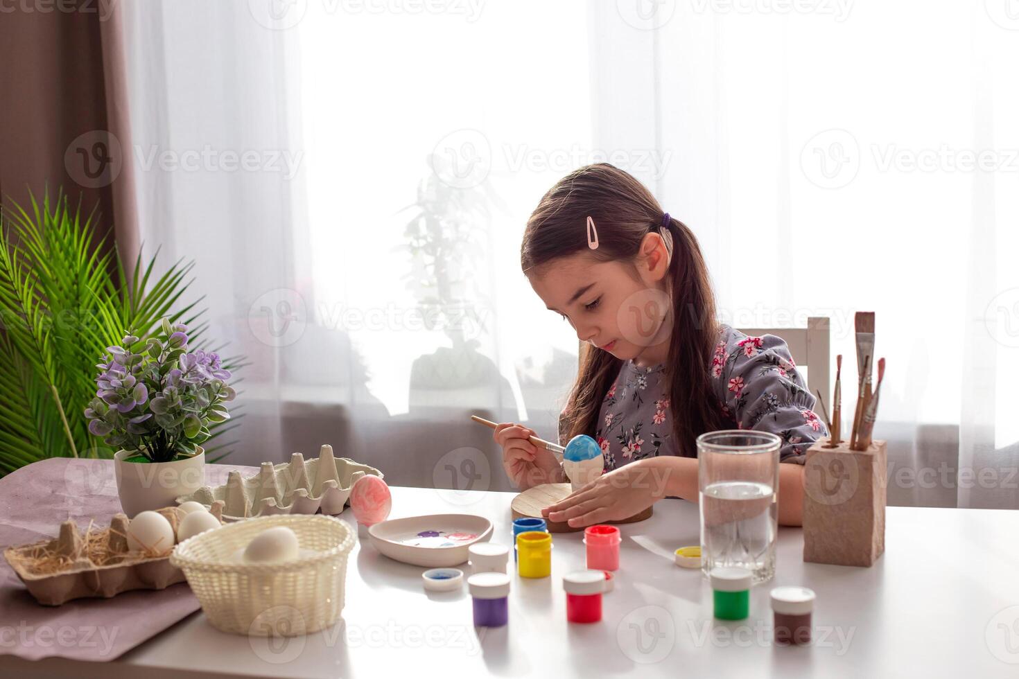 A cute little girl sits at a white table by the window, paints eggs with a brush and paint in jars. photo