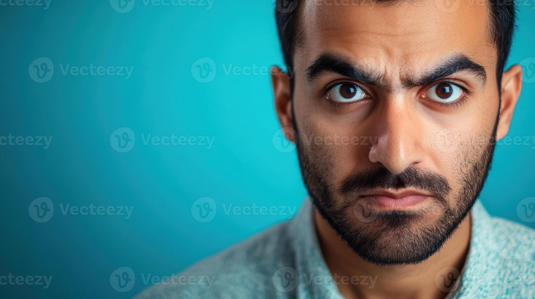 Close-up portrait of an angry man with a deeply furrowed brow and intense expression, glaring directly at the camera, set against a blue background. photo