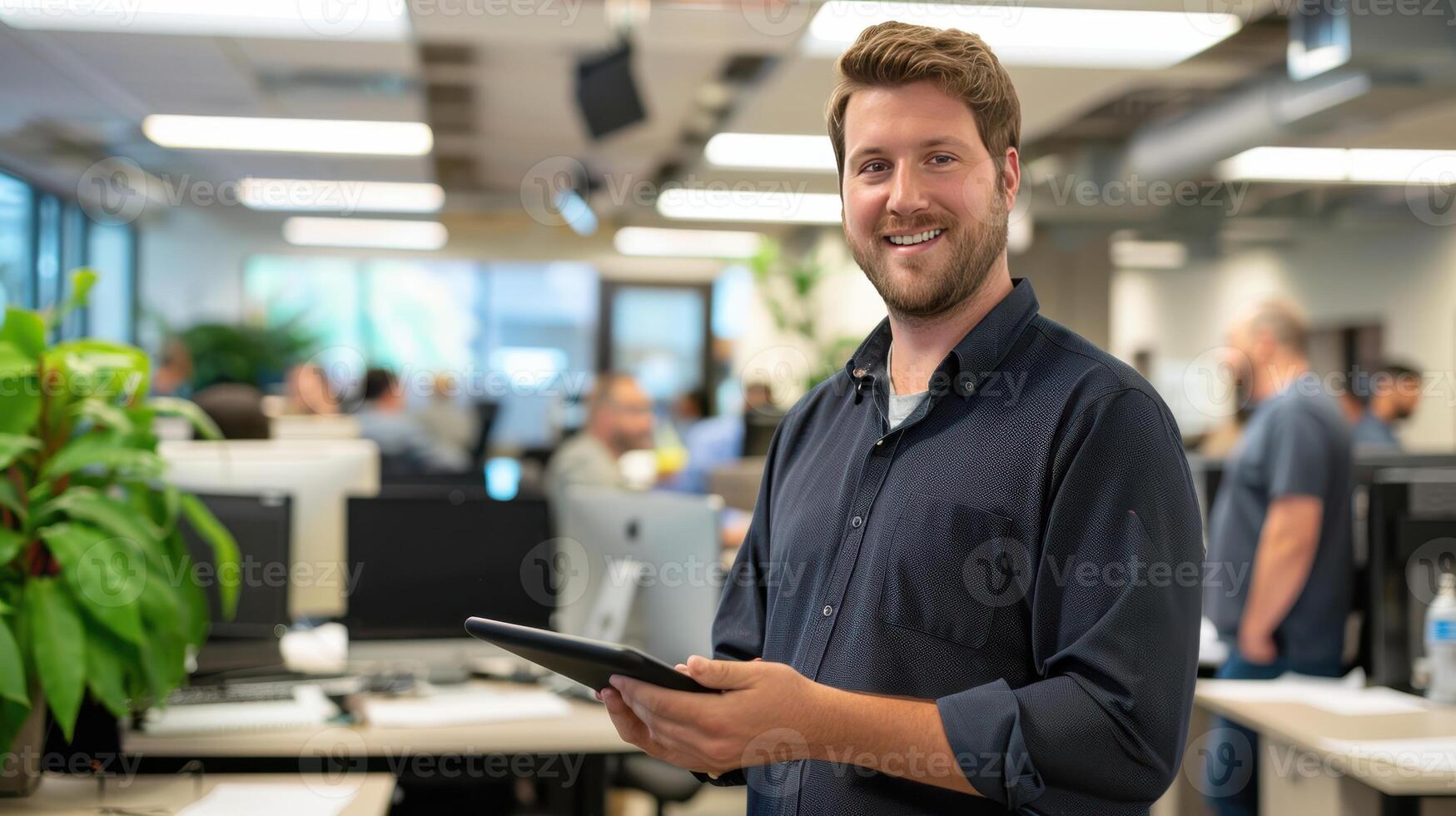 A Young Man Smiles Brightly In A Busy Office Setting While Holding A Tablet photo