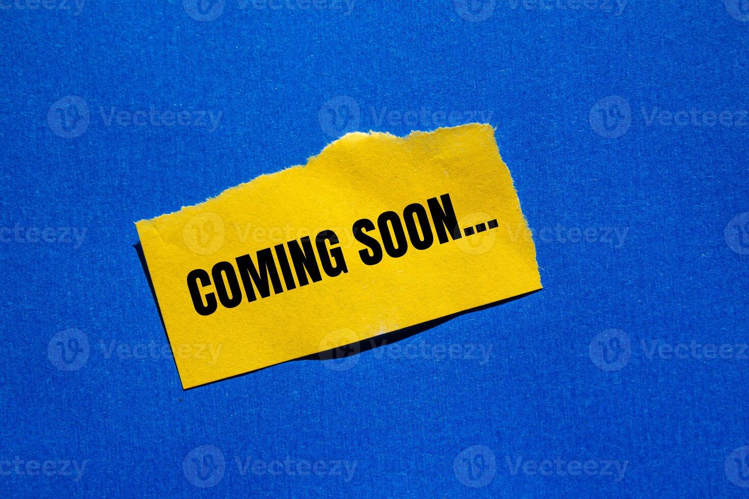 Coming soon message written on ripped torn yellow paper piece with bluebackground. Conceptual coming soon symbol. Copy space. photo