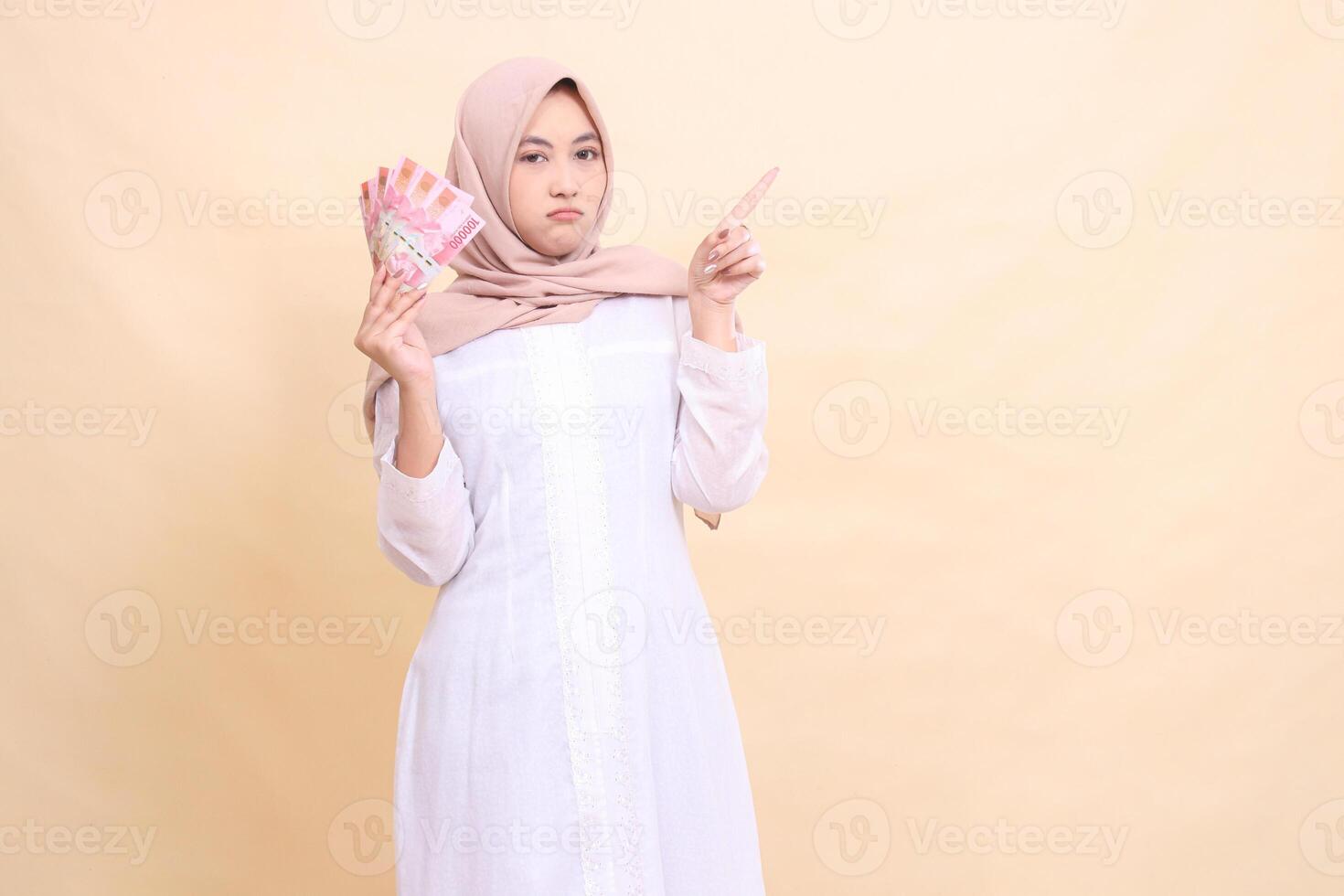 Asian Muslim woman in hijab frowning holding rupiah cash in her hand and upper left. Lifestyle, religious, business and finance concepts photo