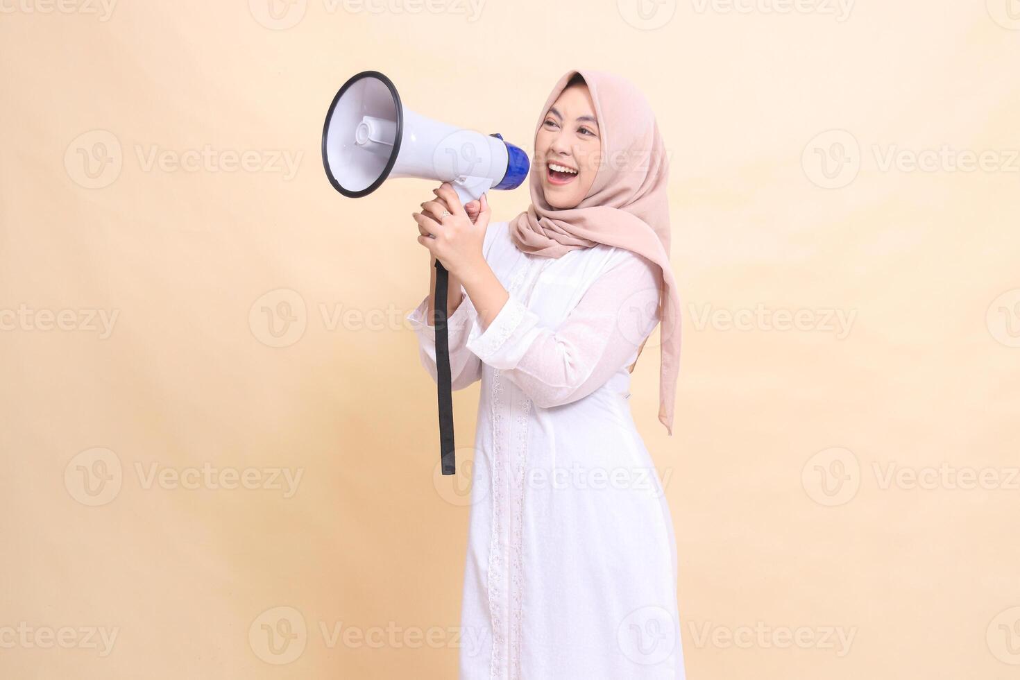 woman asian young wearing hijab slanted to the right candidly shouts cheerfully holding a loudspeaker megaphone with both hands. Technology, broadcast and promotion concept photo