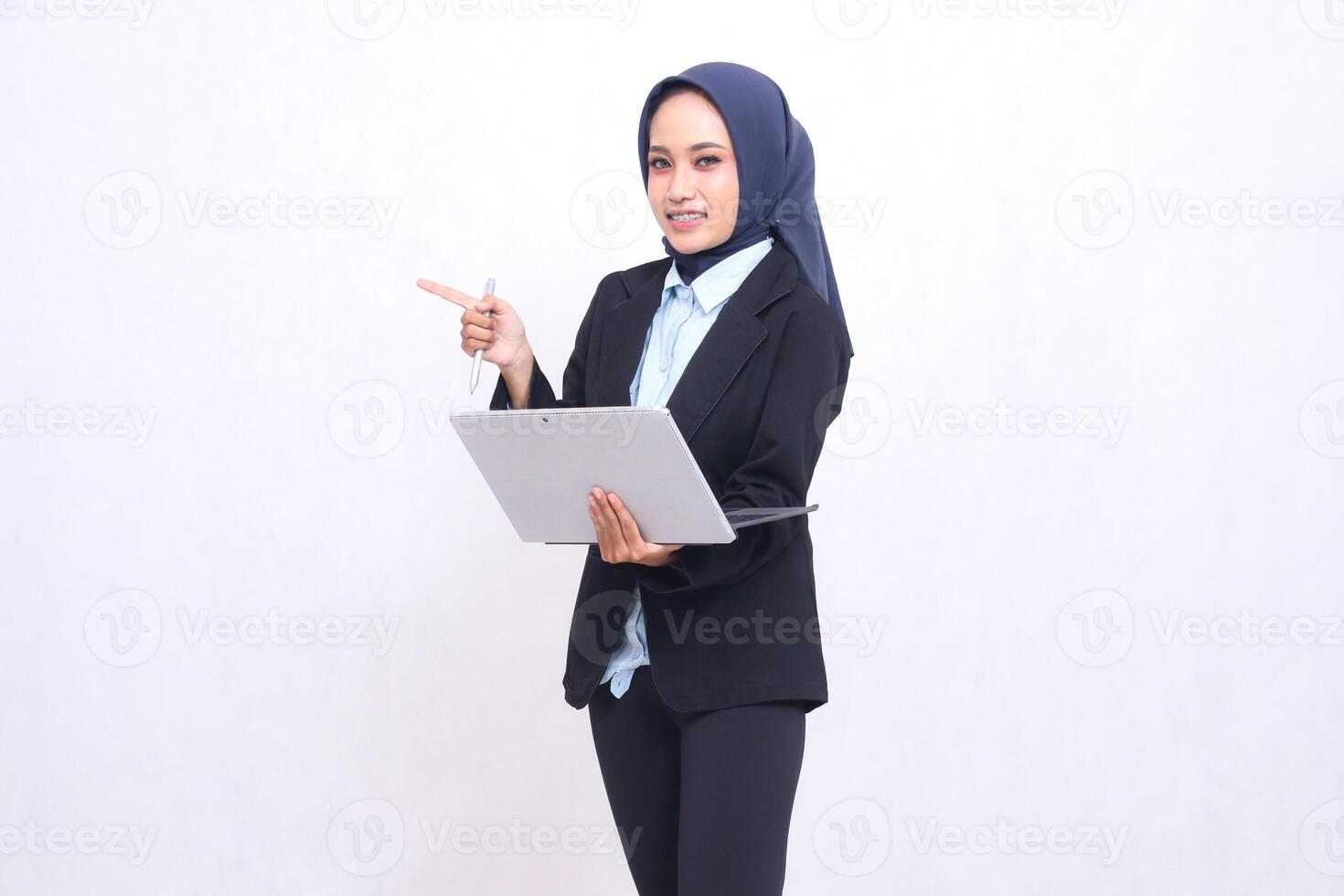 Beautiful Asian office woman wearing a hijab stands cheerfully at the camera gesturing right and operating a tablet laptop. adult Muslim women in blue shirts photo