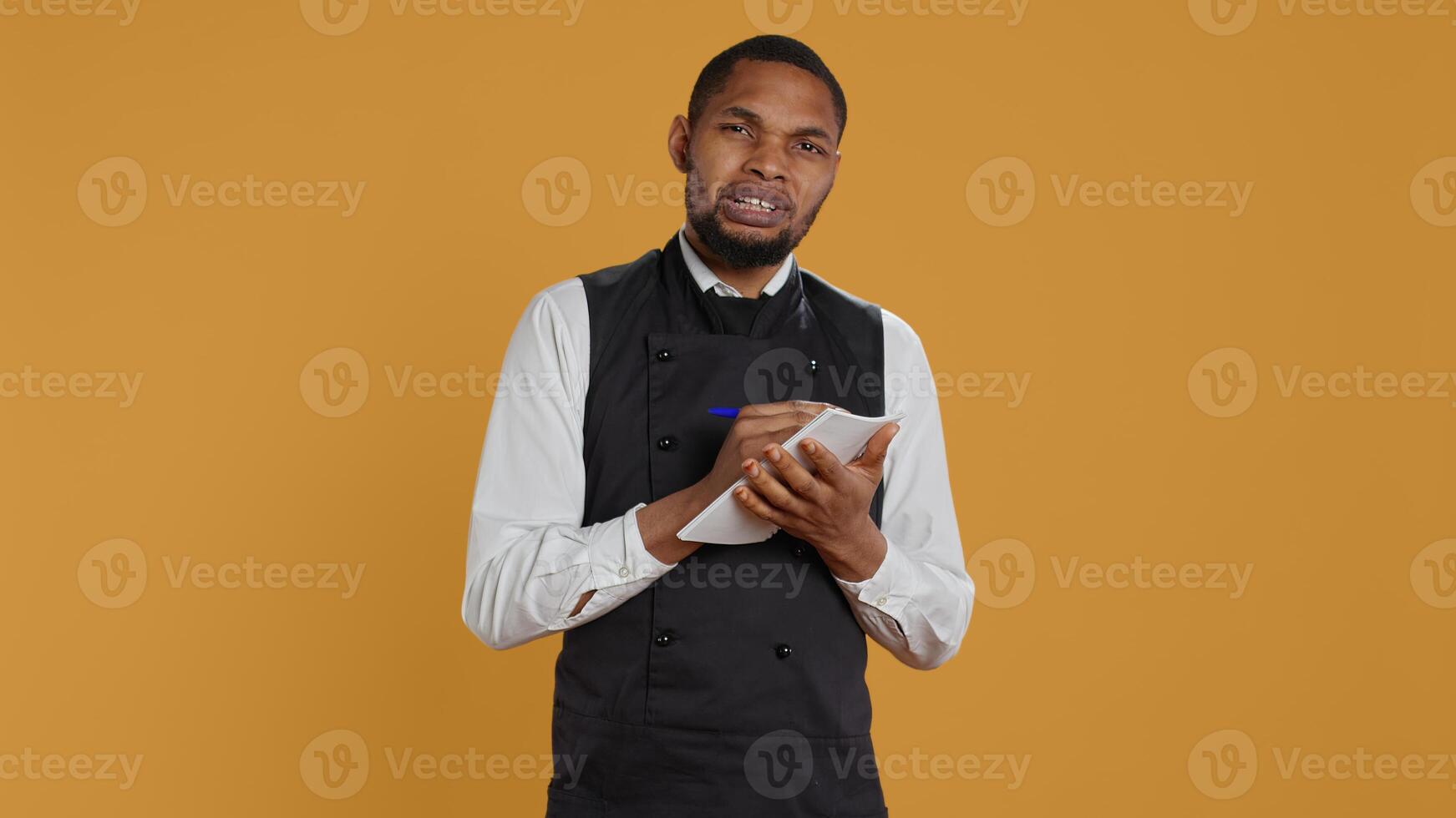 Professional caterer writing clients orders on notepad for the bill, takes notes of desired food against yellow background. Waiter with apron, notebook service for customer satisfaction. Camera B. photo