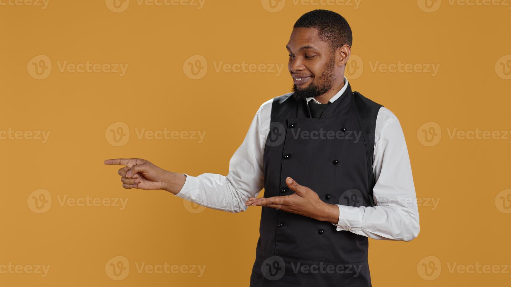 Waiter in professional uniform pointing at something to the left or right, creating a commercial ad for five star service oriented restaurant. Catering staff in hospitality industry. Camera B. photo